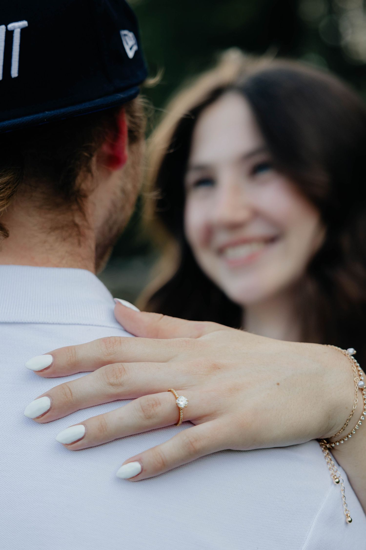 Close-up of engaged woman showing pearl ring with white nails resting on partner's shoulder.