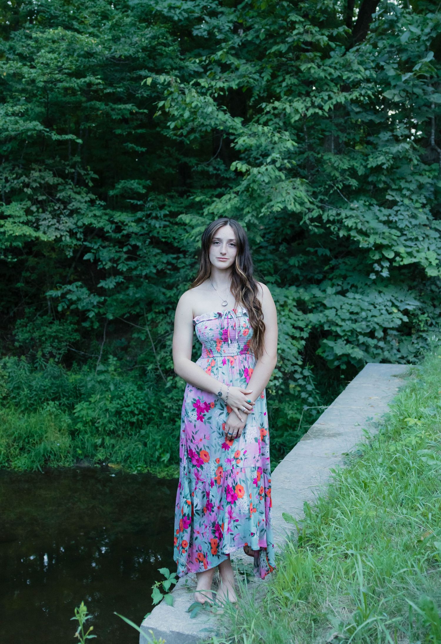 Young woman in strapless floral maxi dress standing on stone ledge beside pond in green forest.