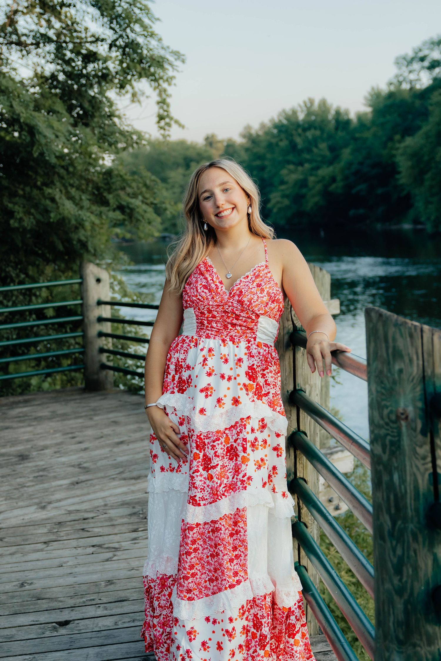 Smiling blonde woman in red floral maxi dress standing on wooden bridge over river at sunset.