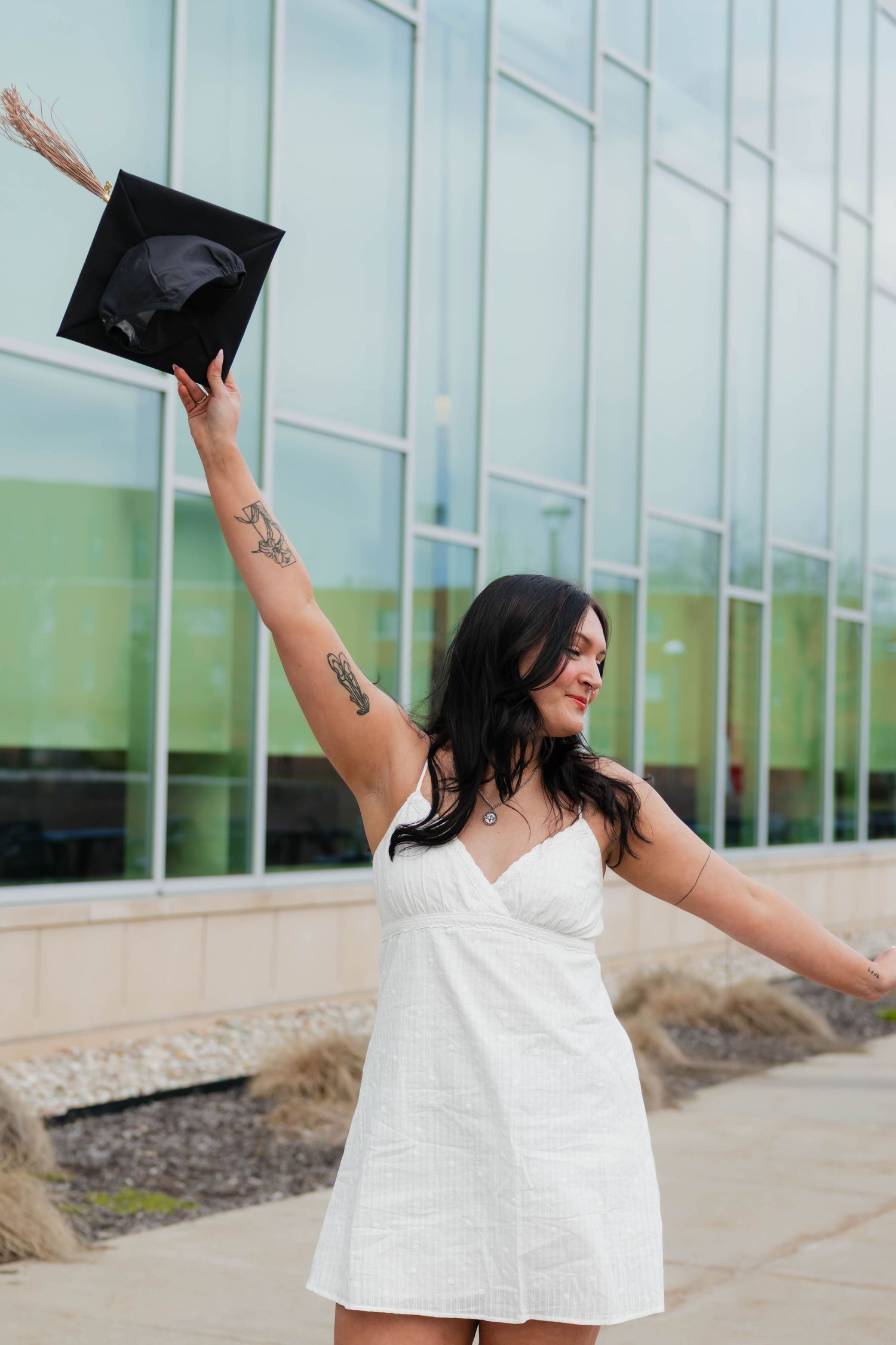 Tattooed brunette graduate in white dress raising black cap triumphantly outside glass building.