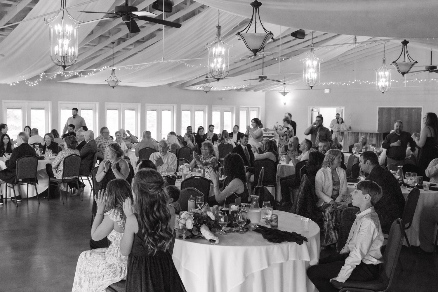 Black and white wide shot of wedding reception hall filled with guests seated at round tables.