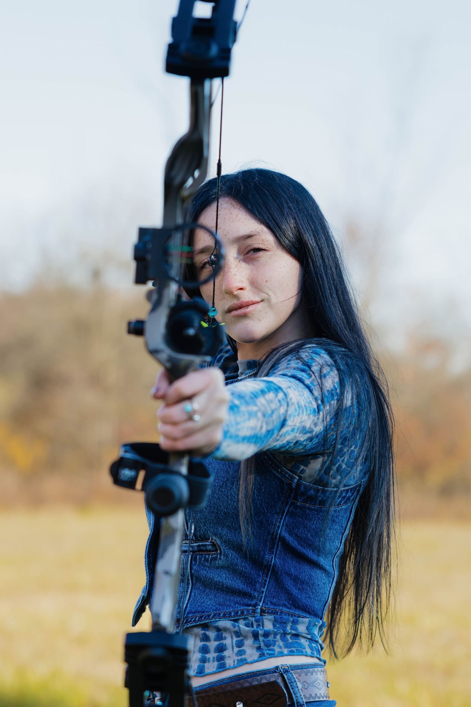 Young woman with long black hair aiming a compound bow outdoors in autumn setting.