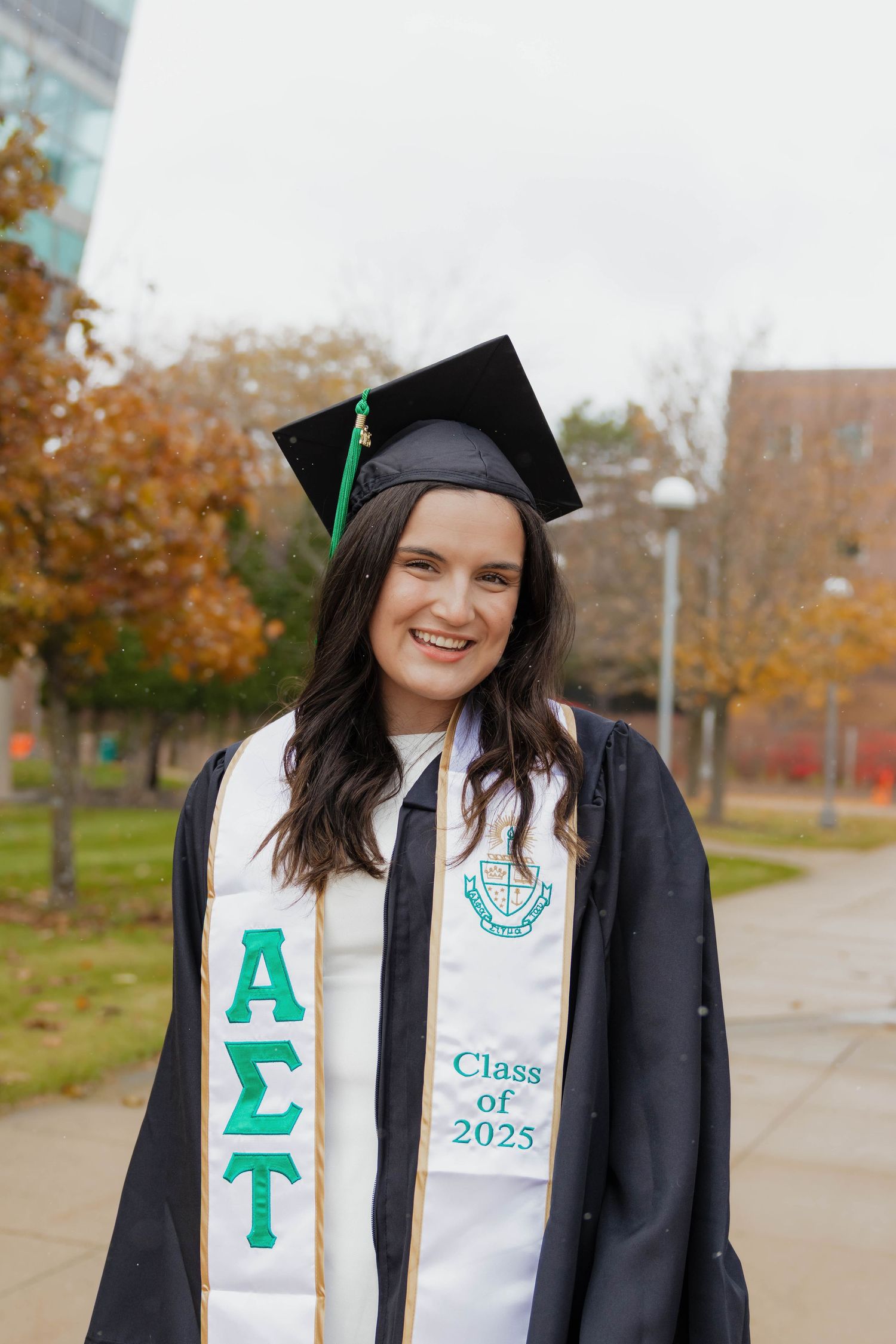 Smiling graduate in cap and gown wearing Alpha Sigma Tau stole Class of 2025 outdoors.