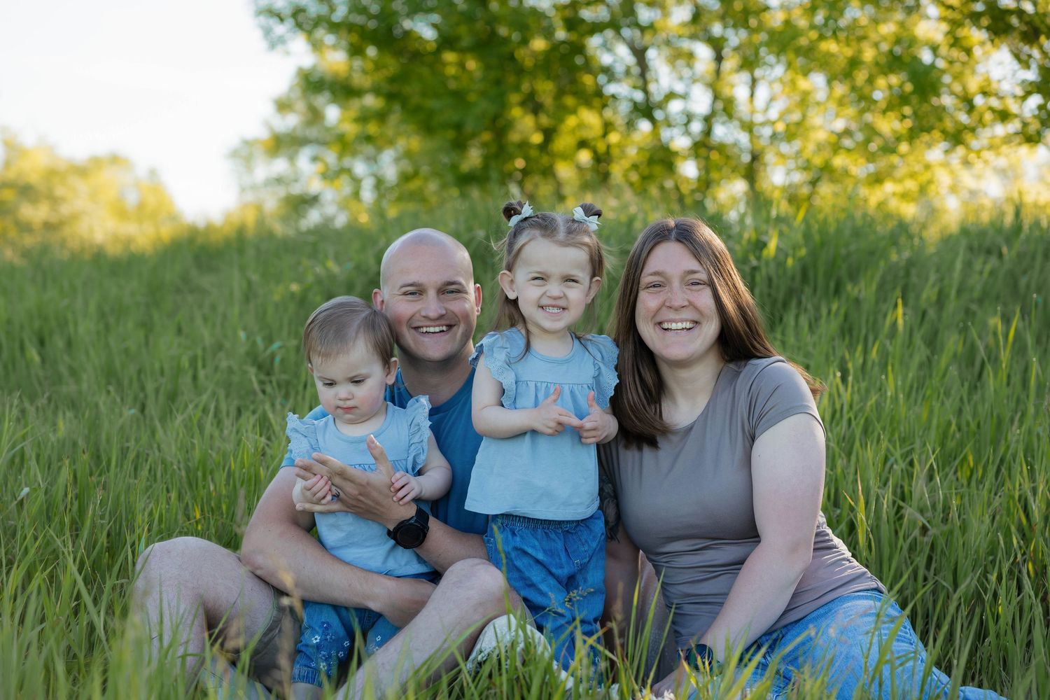 Happy family of four sitting together smiling in a beautiful green grassy field at sunset.