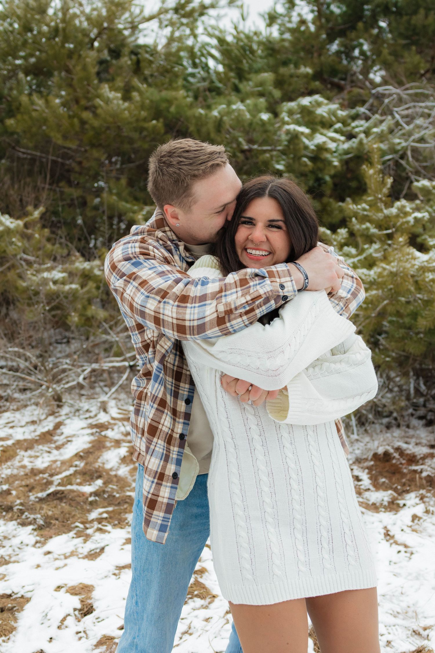 Man hugging woman from behind in snowy forest, both smiling, wearing casual winter outfits outdoors.