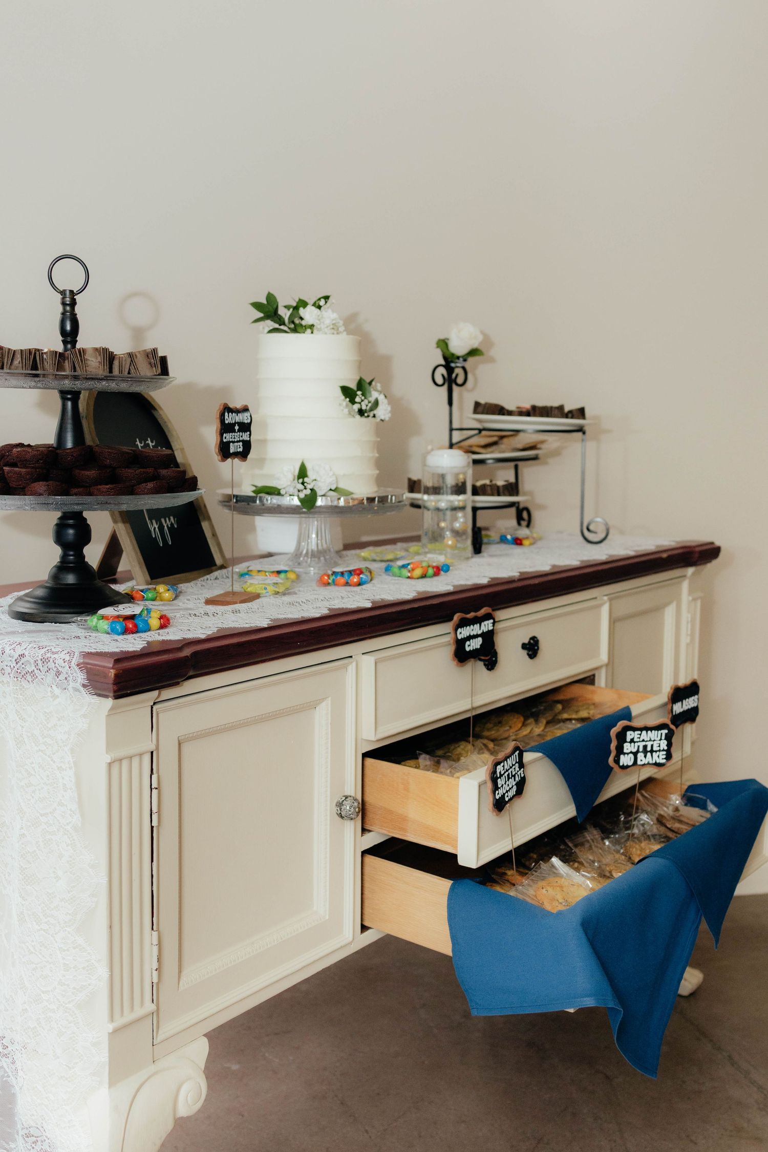 Wedding dessert table with white tiered cake, treats, and open drawer displaying labeled snacks.