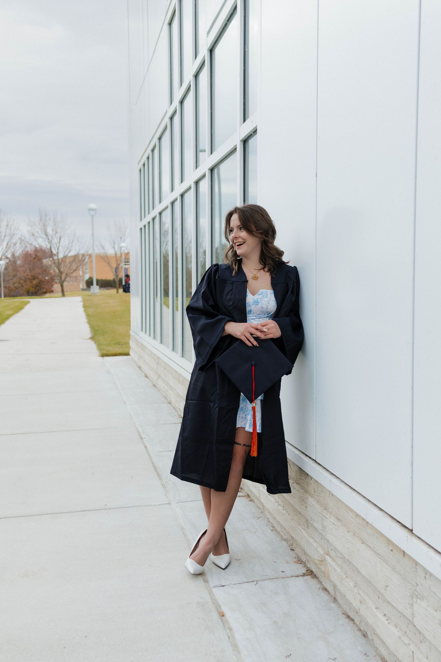 Young woman in black graduation gown leans against glass building wall holding her cap while looking away.