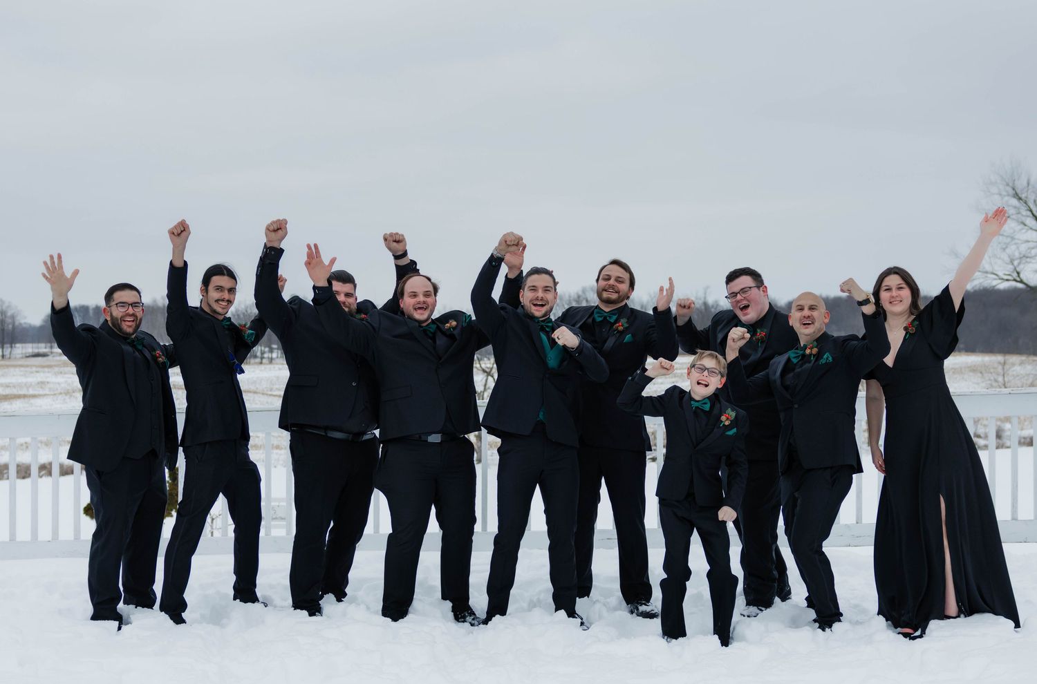 Groomsmen and wedding party in black attire cheering with raised arms in snowy outdoor setting.
