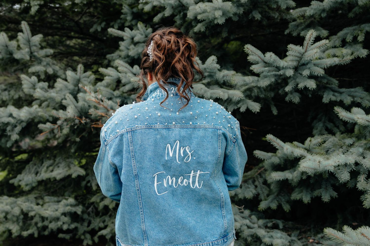 Woman wearing a personalized denim jacket with Mrs. Emcott written on the back, standing in front of evergreen trees.
