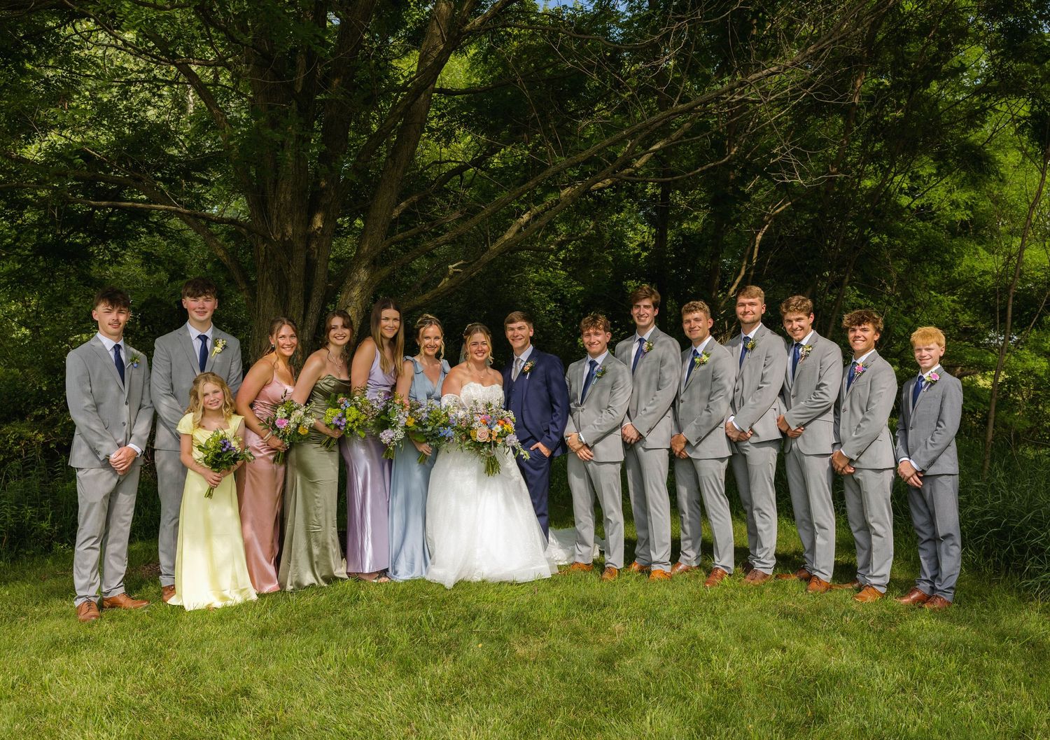 Wedding party posing outdoors in lush green garden, bridesmaids in pastel gowns and groomsmen in gray suits flanking bride and groom.