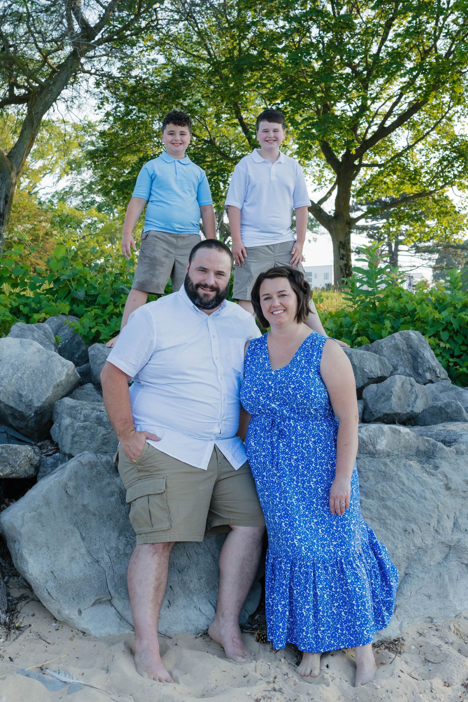 Family of four posing on rocky boulders surrounded by green trees at the beach.
