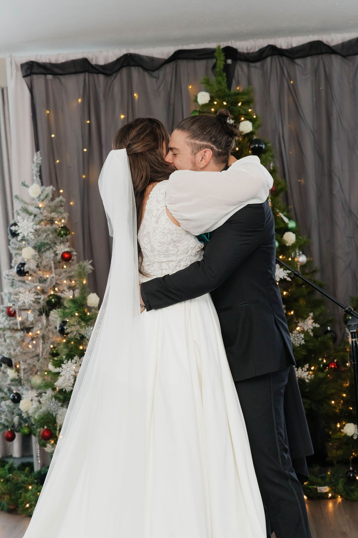 Bride and groom embracing in first kiss during winter wedding ceremony with Christmas trees behind.