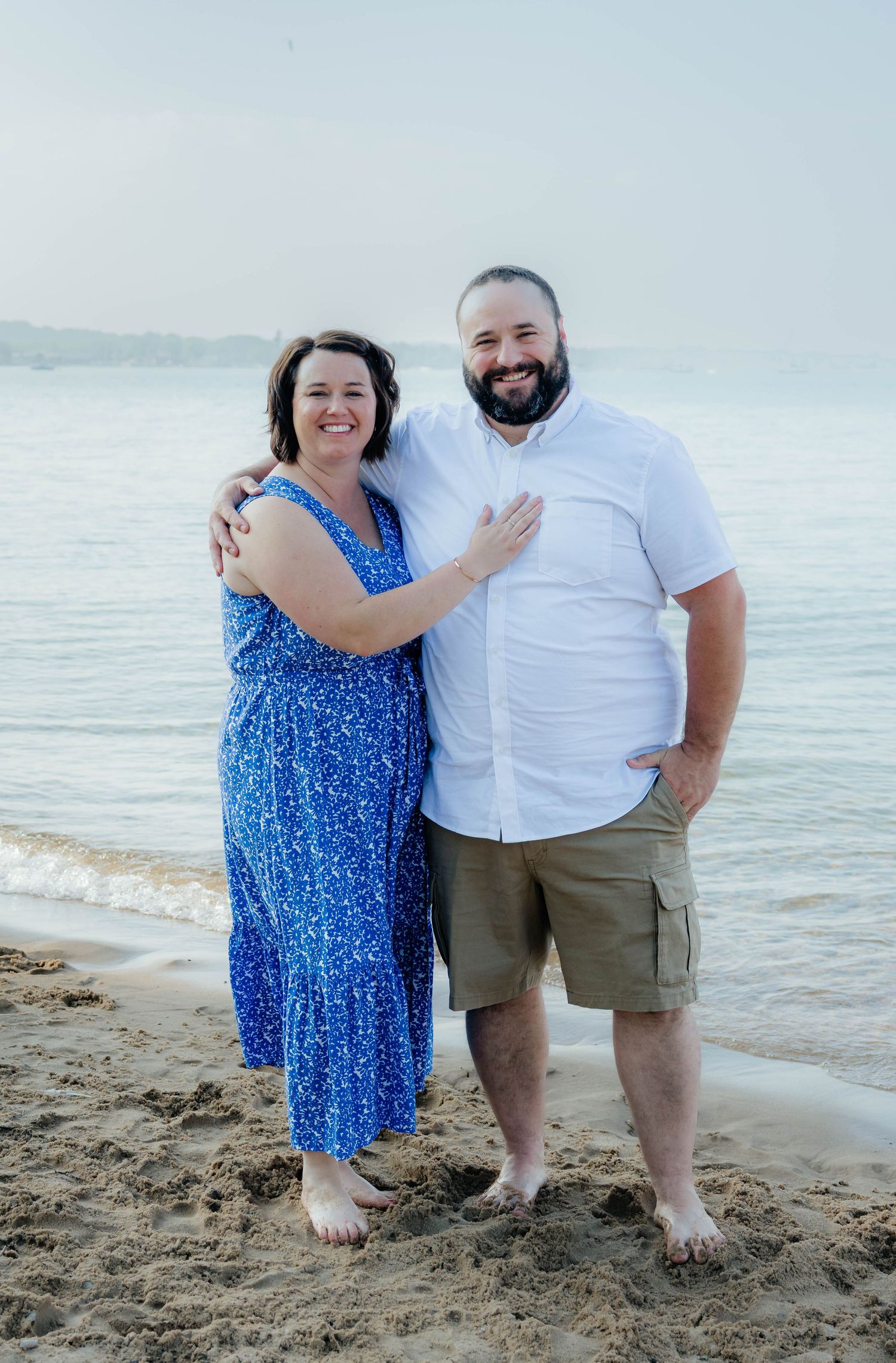Smiling couple embracing on a sandy beach with calm ocean waves in the background.