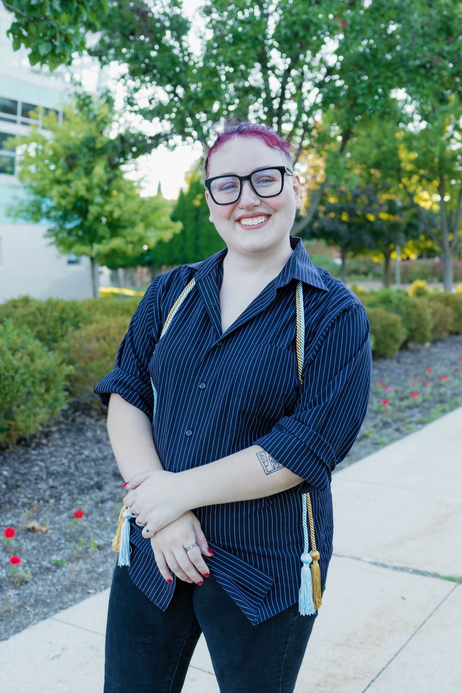 Graduate with pink hair smiling outdoors in striped shirt with gold and blue tassels.