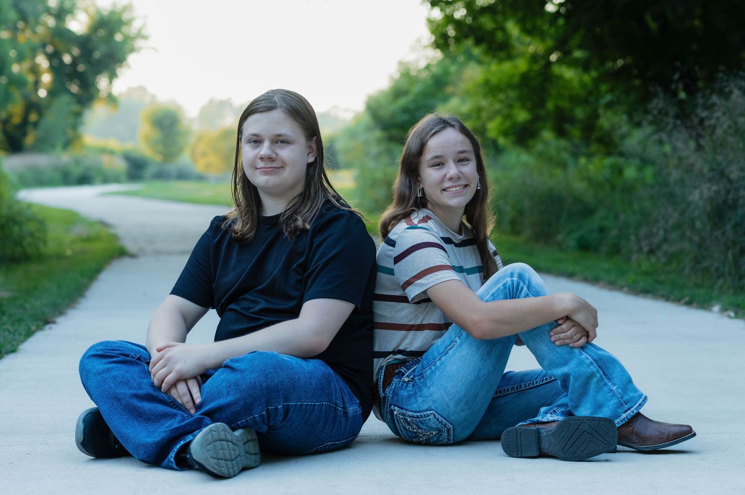 Two teenagers sitting back-to-back on a path outdoors, smiling, surrounded by green trees.