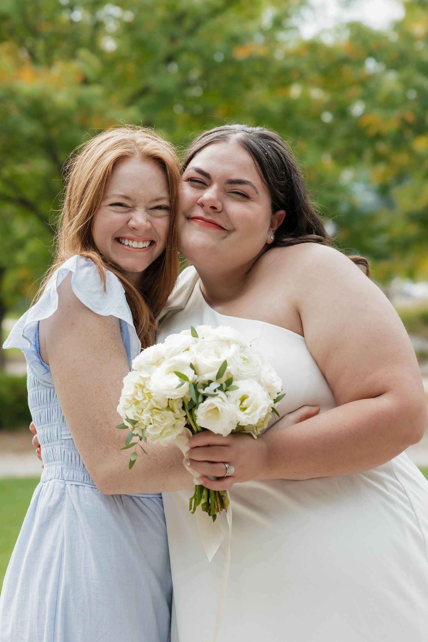 Bride in white dress holding white bouquet shares joyful hug with redhead friend outdoors.