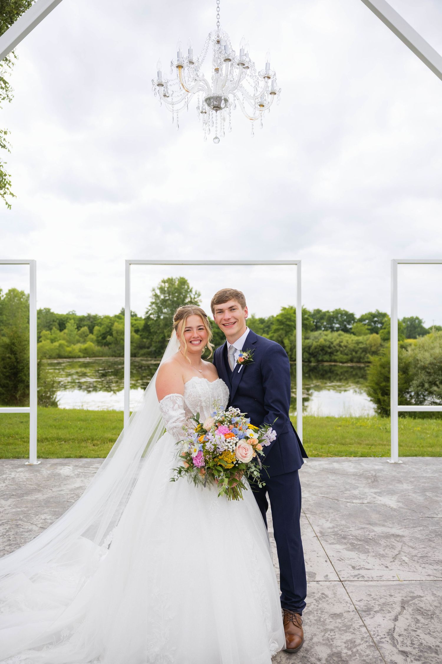 Bride and groom smiling under crystal chandelier at outdoor wedding ceremony with white arch frames and pond backdrop.