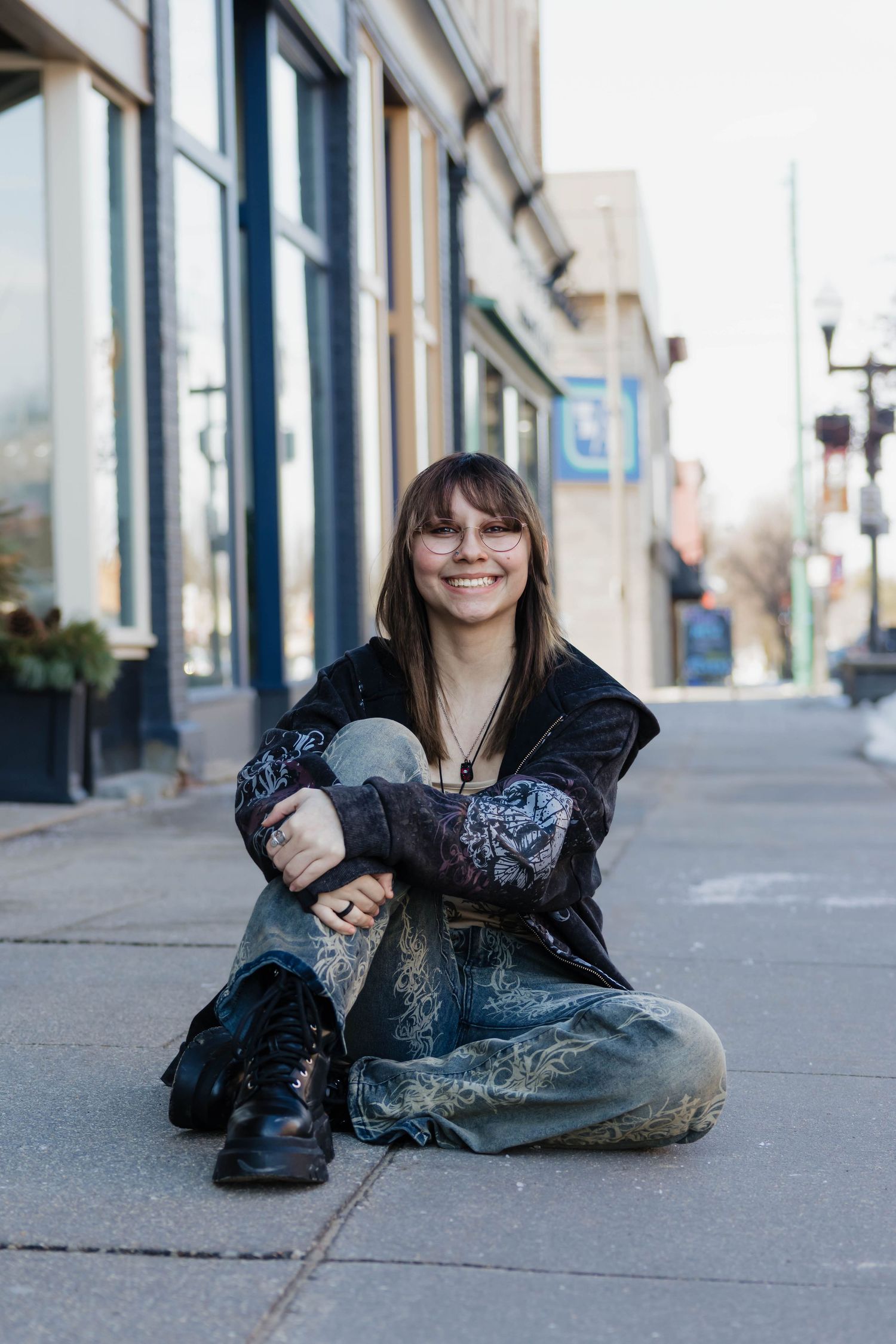 Young woman sitting cross-legged on urban sidewalk wearing decorated jeans and black boots, smiling outdoors.