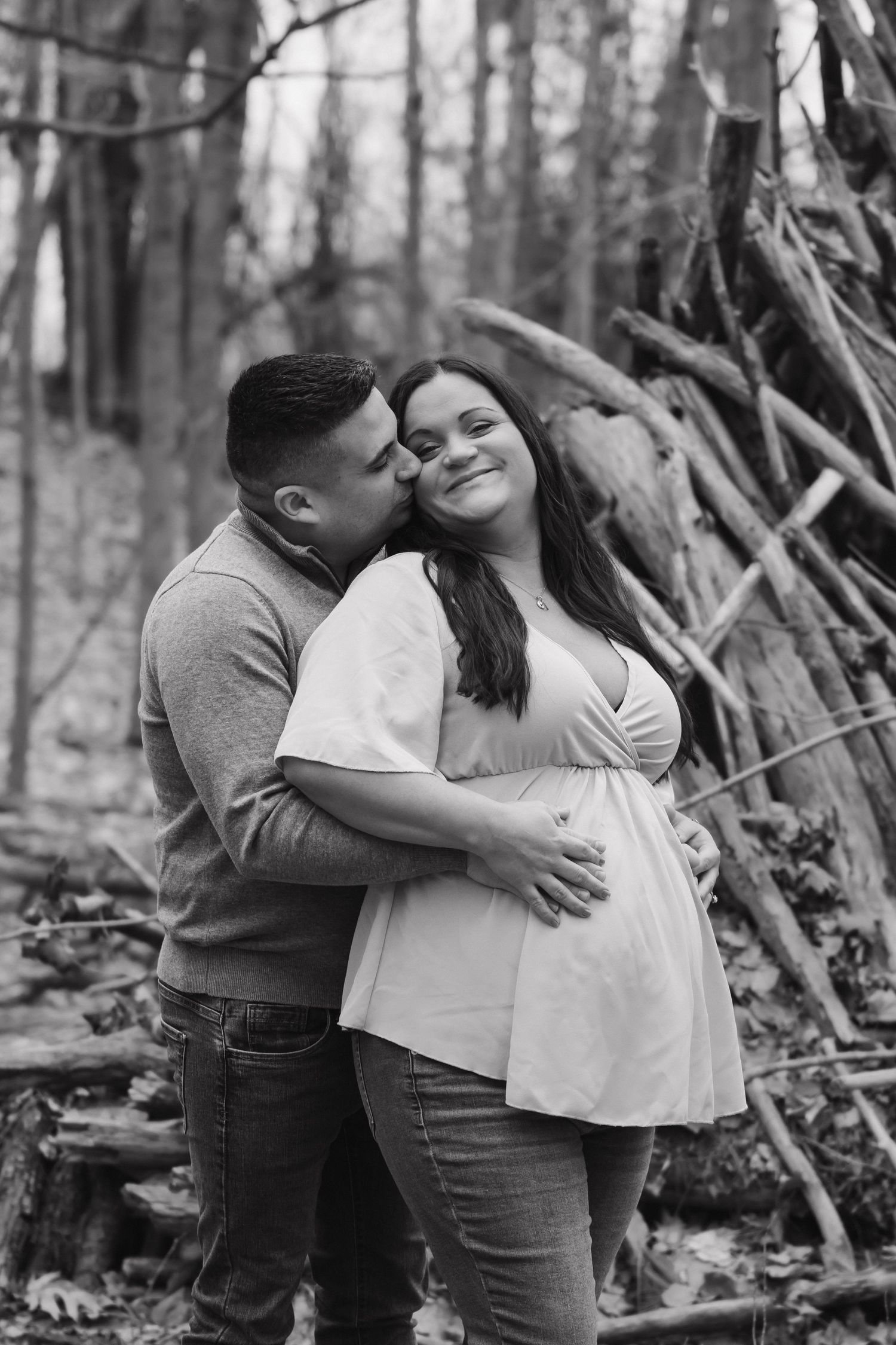Black and white photo of a couple sharing a tender moment, man kissing pregnant woman's cheek in woods.