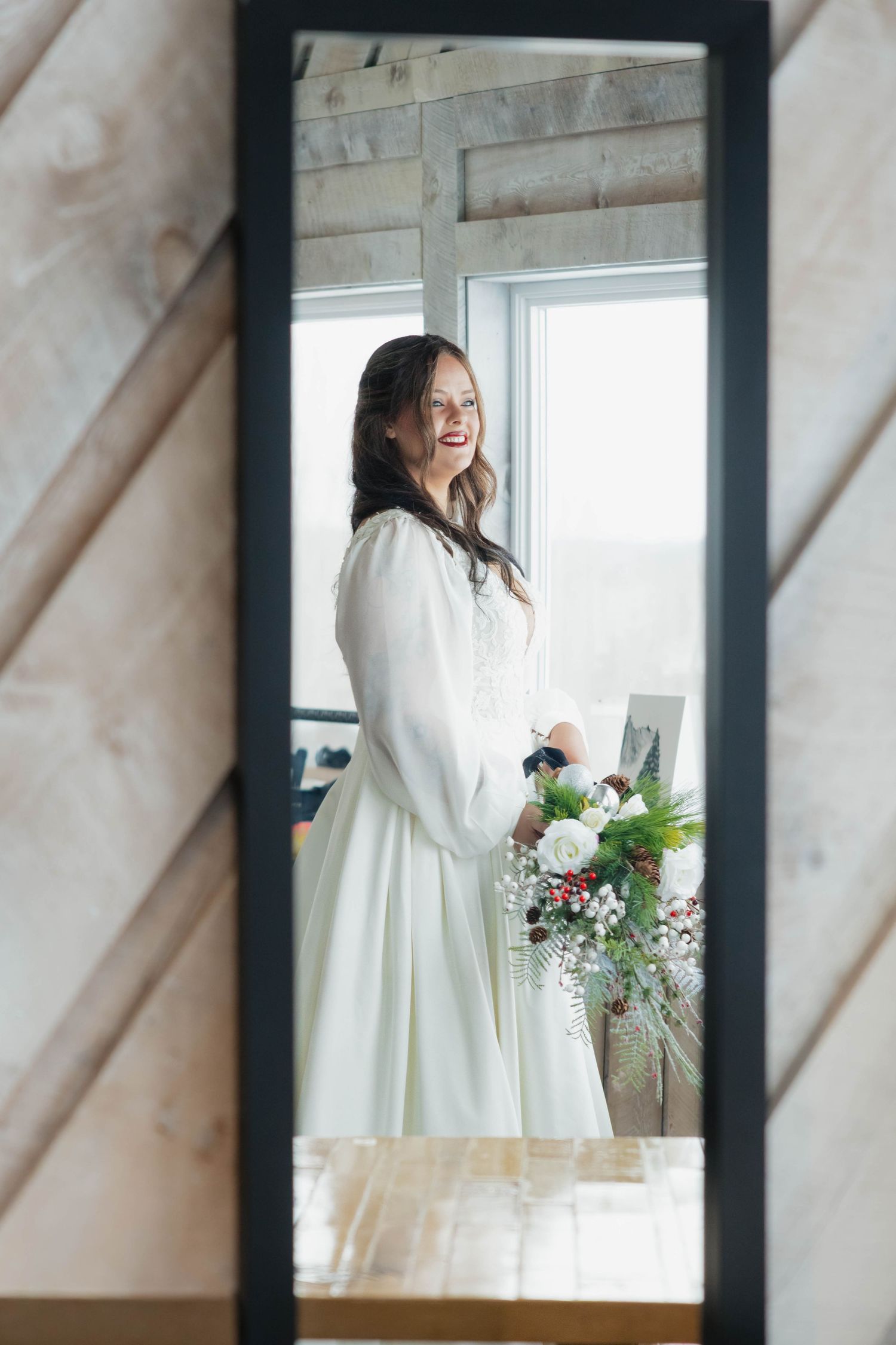 Smiling bride in white long-sleeve wedding dress holding bouquet reflected in a full-length mirror.