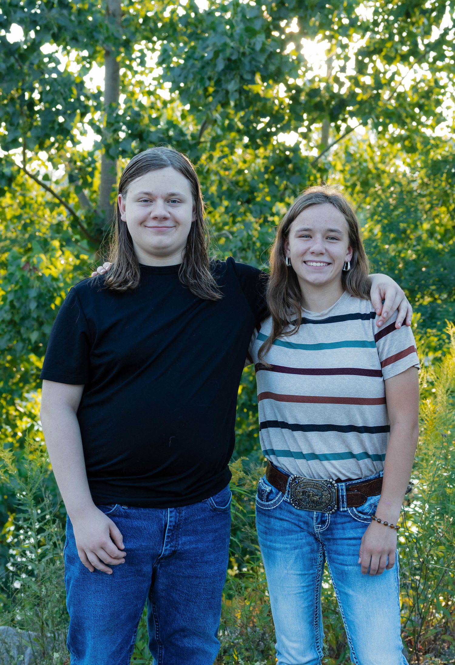 Two smiling teenagers pose together outdoors in golden evening light surrounded by green trees.