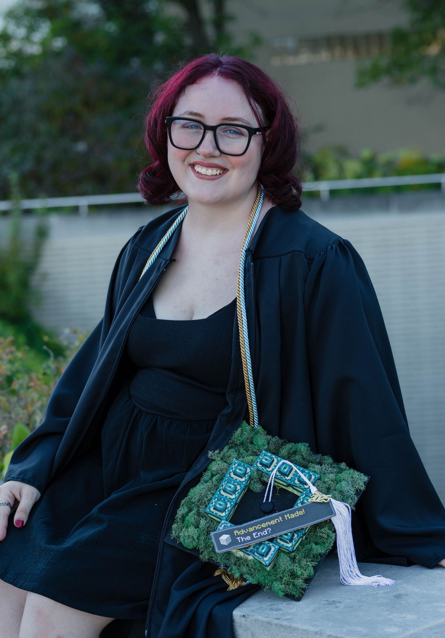 Smiling graduate in black gown holding decorated graduation cap with moss design outdoors.