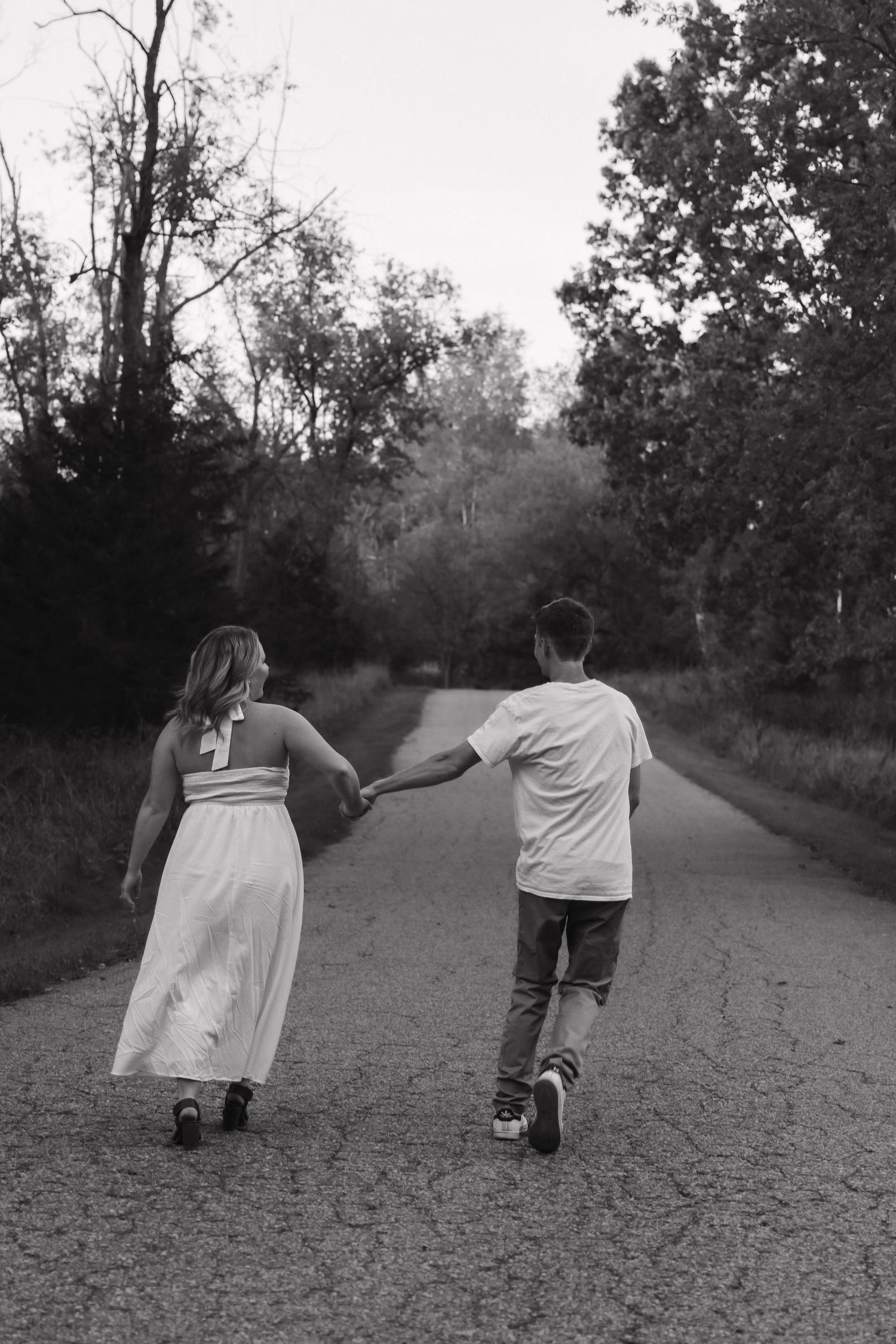 Black and white photo of couple holding hands walking down a tree-lined road.