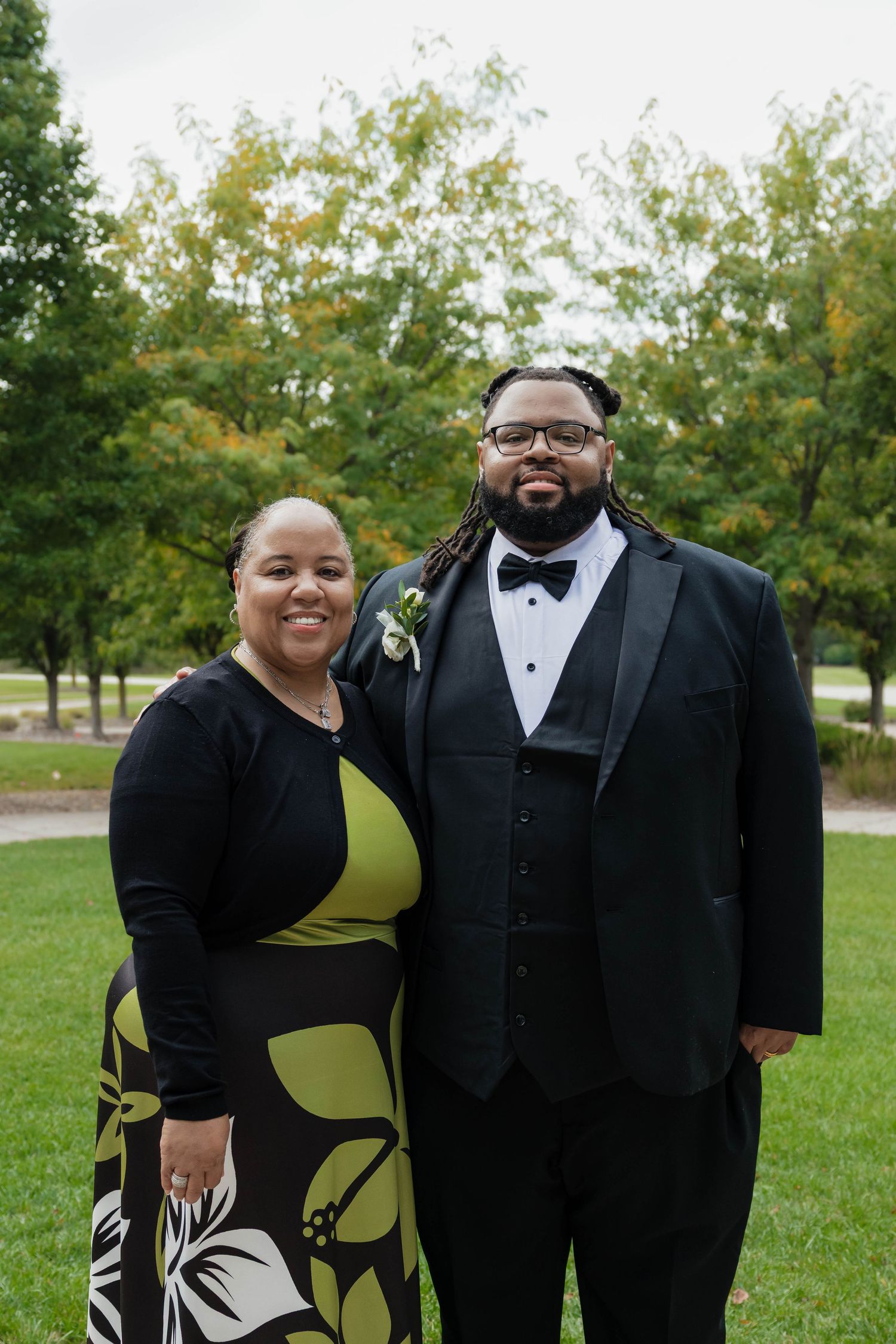 Smiling couple poses outdoors, man in tuxedo with boutonniere and woman in floral dress.