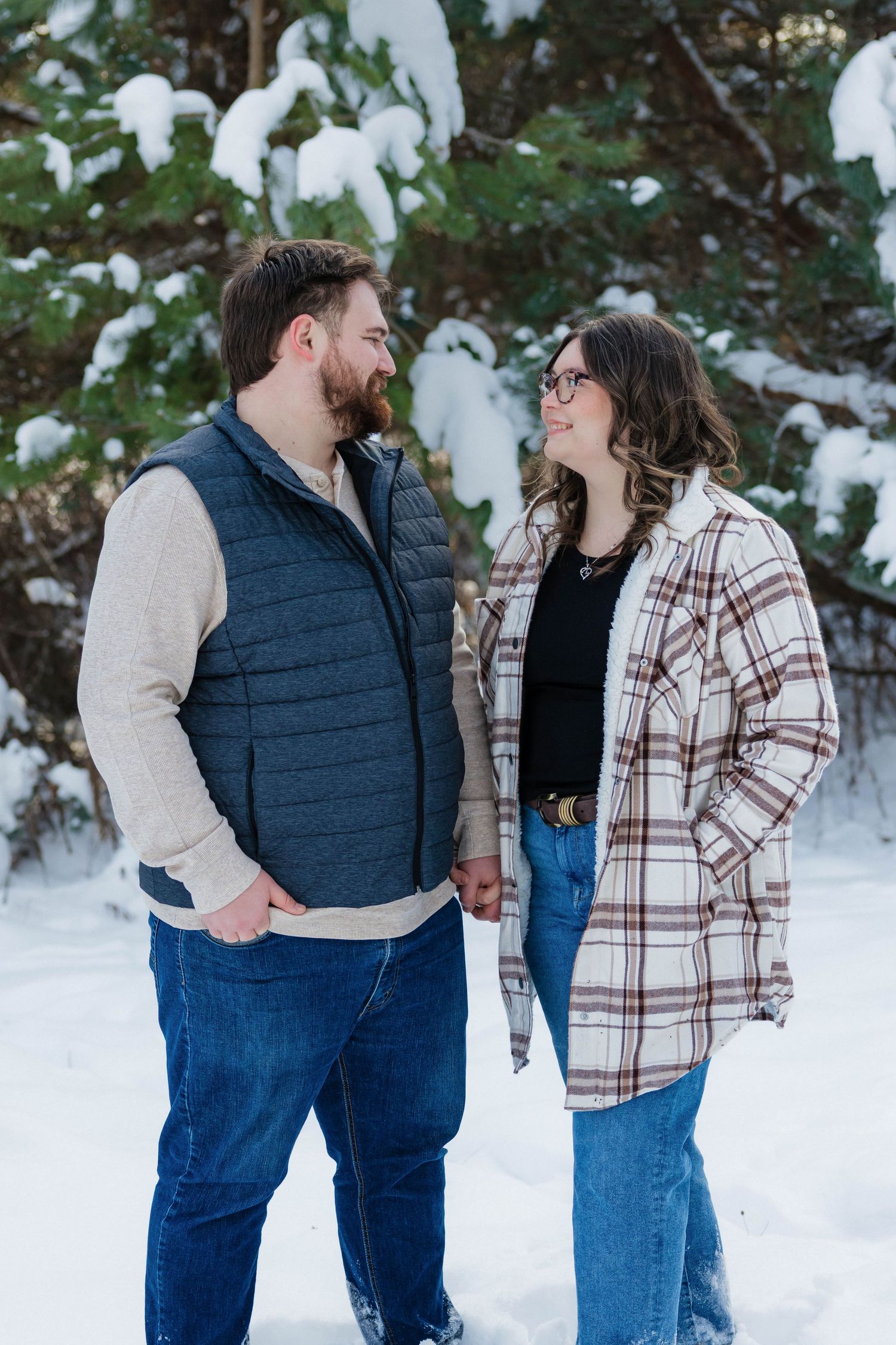 Couple holding hands and smiling at each other in a snowy winter setting with snow-covered pine trees.