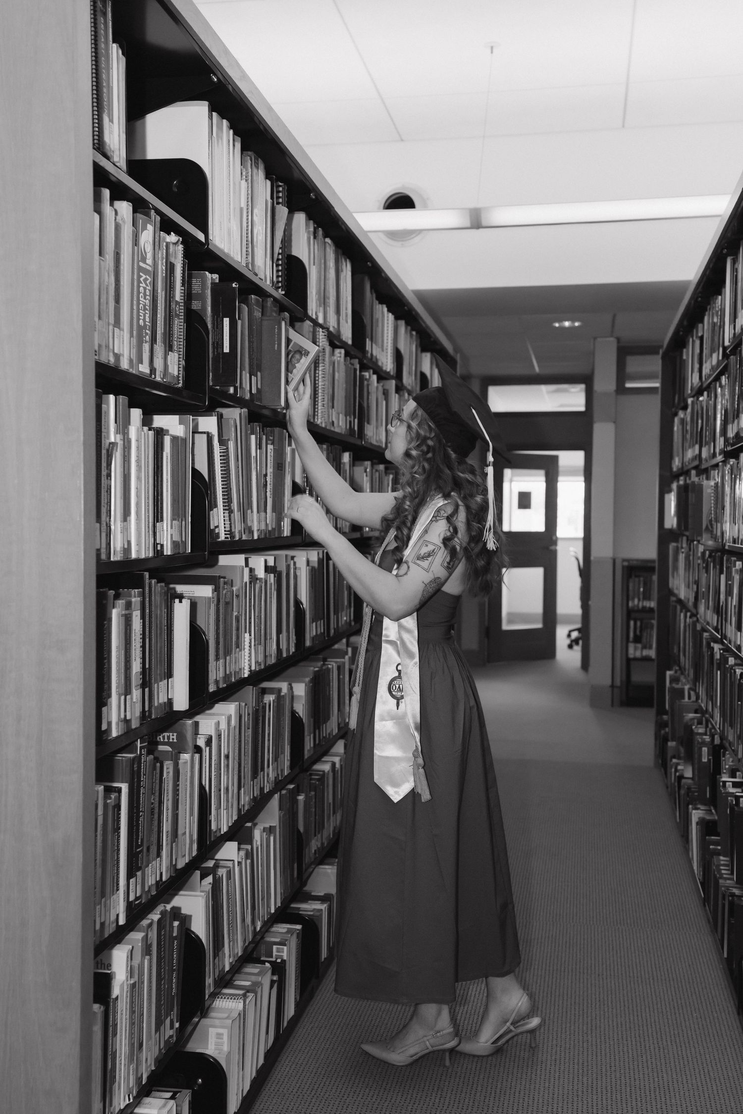 Black and white photo of a graduate in a dress browsing books along a library bookshelf aisle.