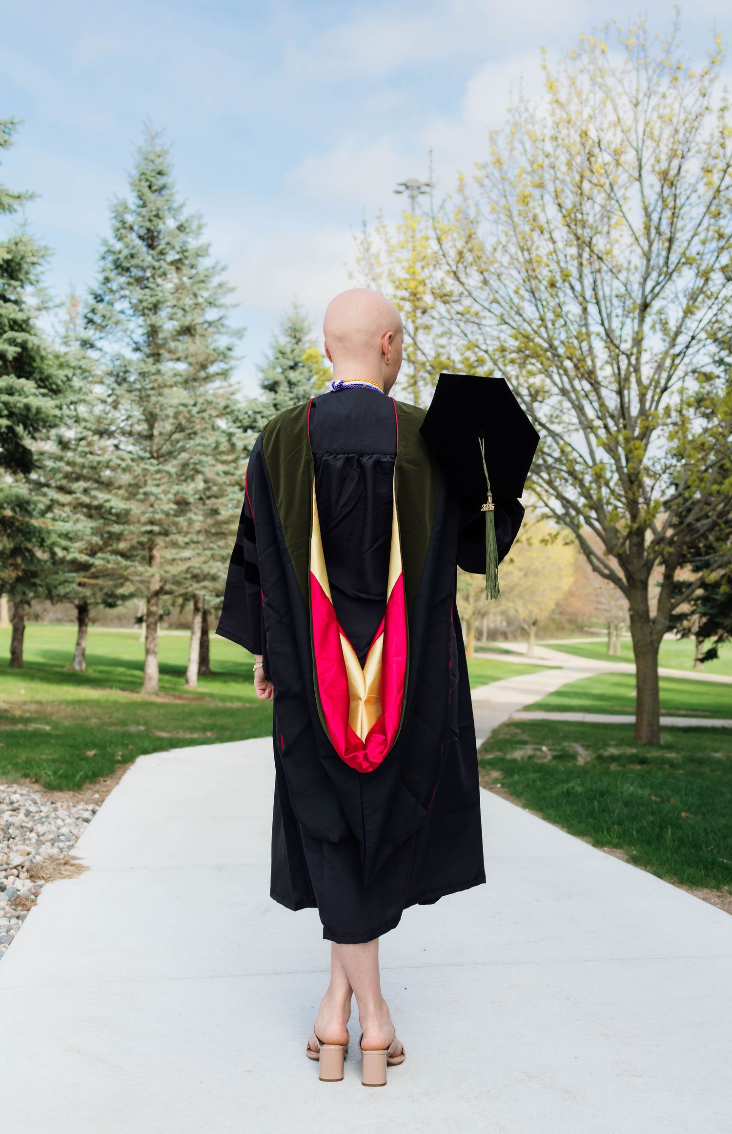 Bald graduate in full doctoral regalia with red and gold hood walking along campus pathway.