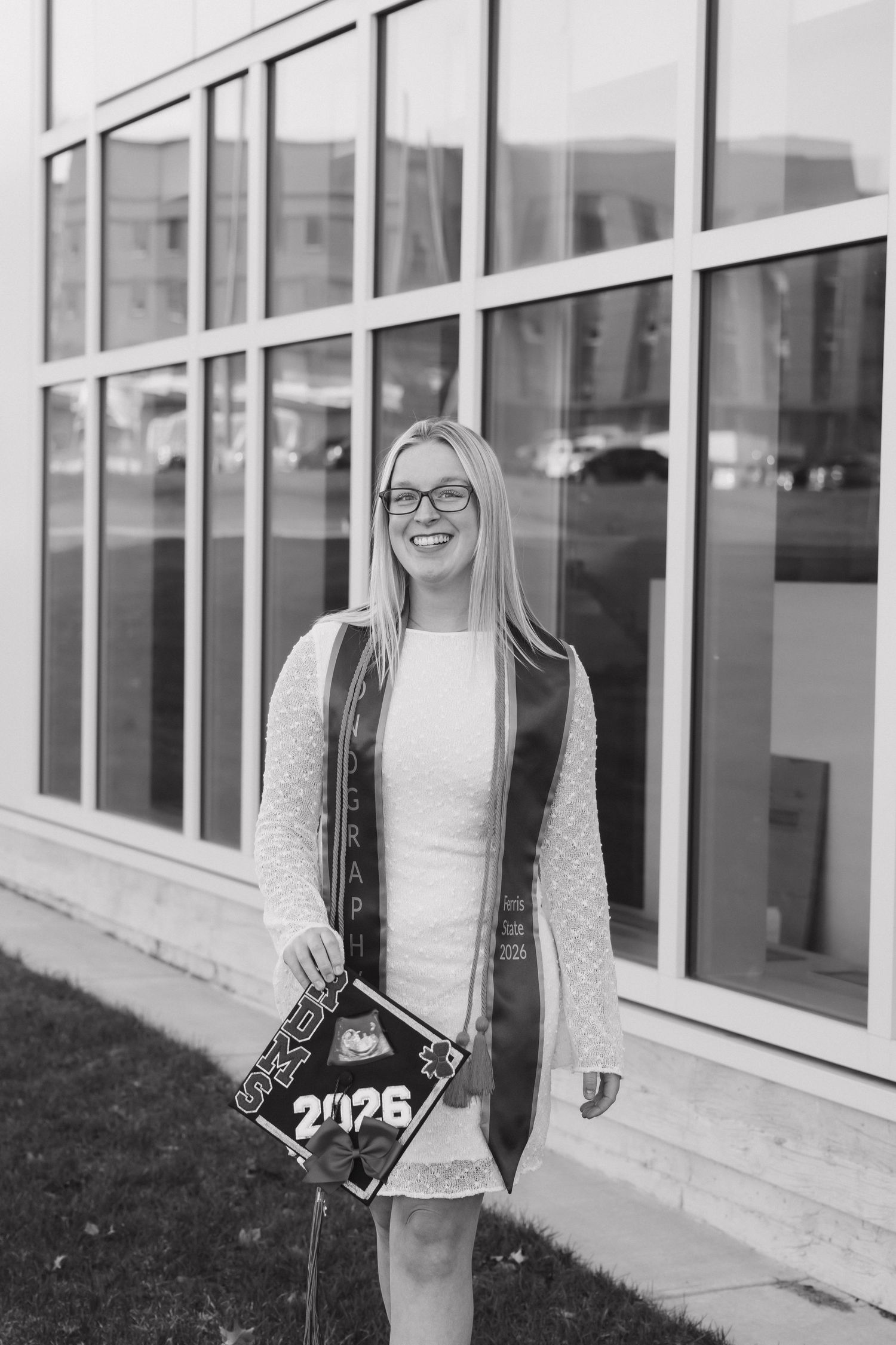 Black and white photo of smiling female graduate holding 2026 decorated cap outside campus building.