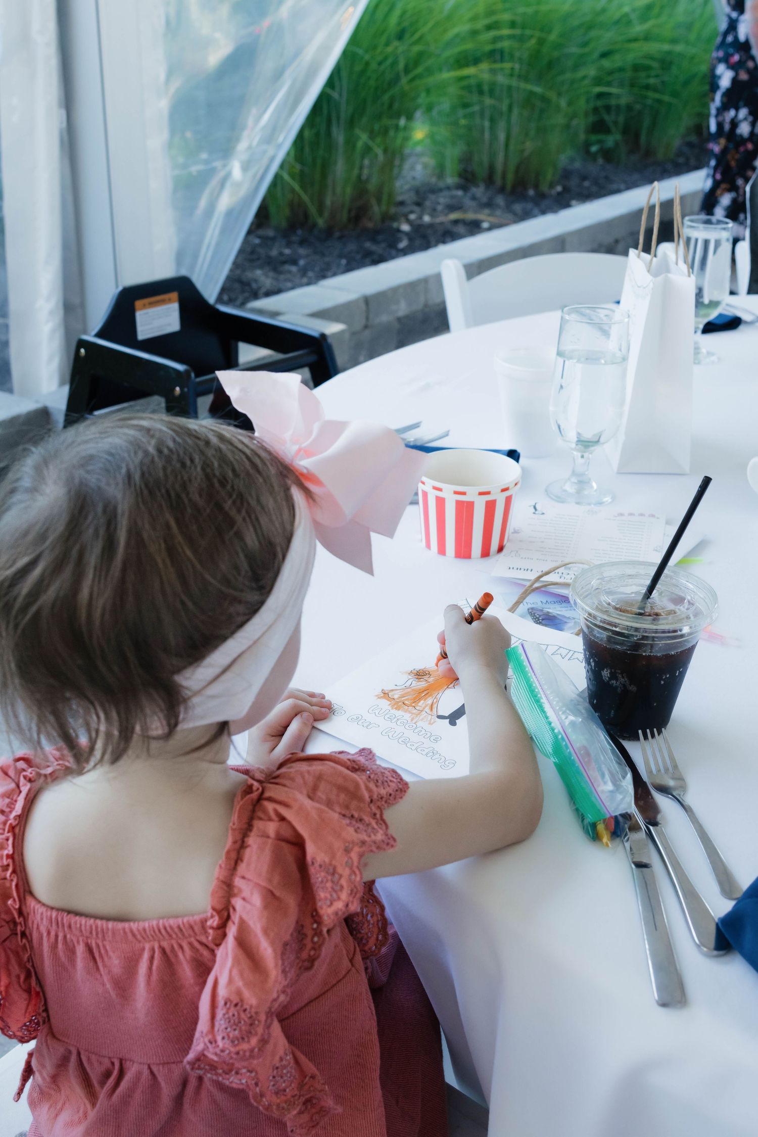 Young girl with pink bow colors activity book at wedding reception table with drinks nearby.