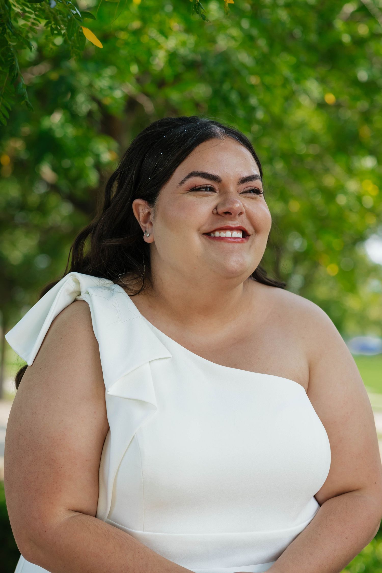 Smiling bride in a white one-shoulder gown standing outdoors under green trees.