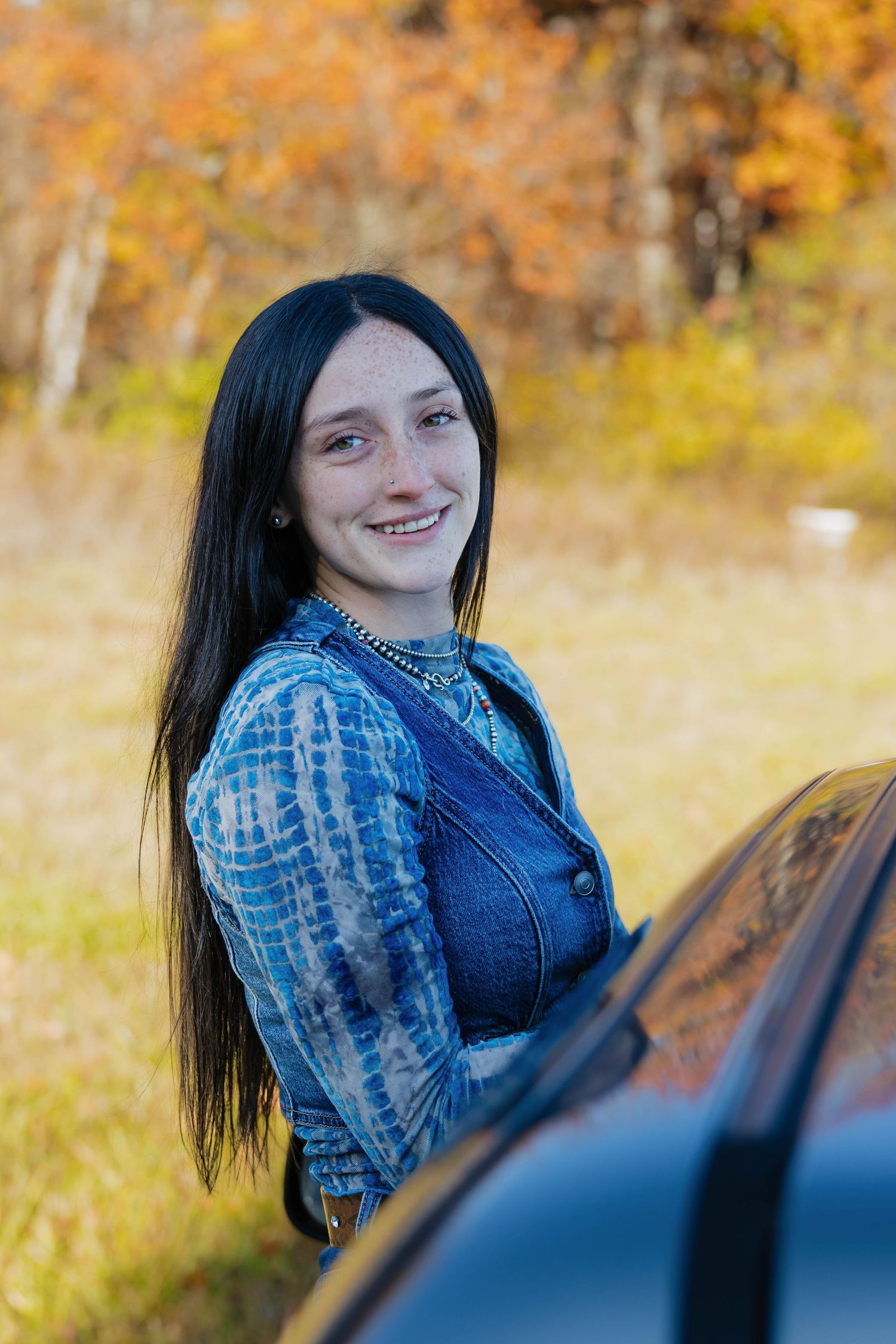 Smiling teenage girl with dark hair and freckles standing by car in autumn setting wearing denim outfit.