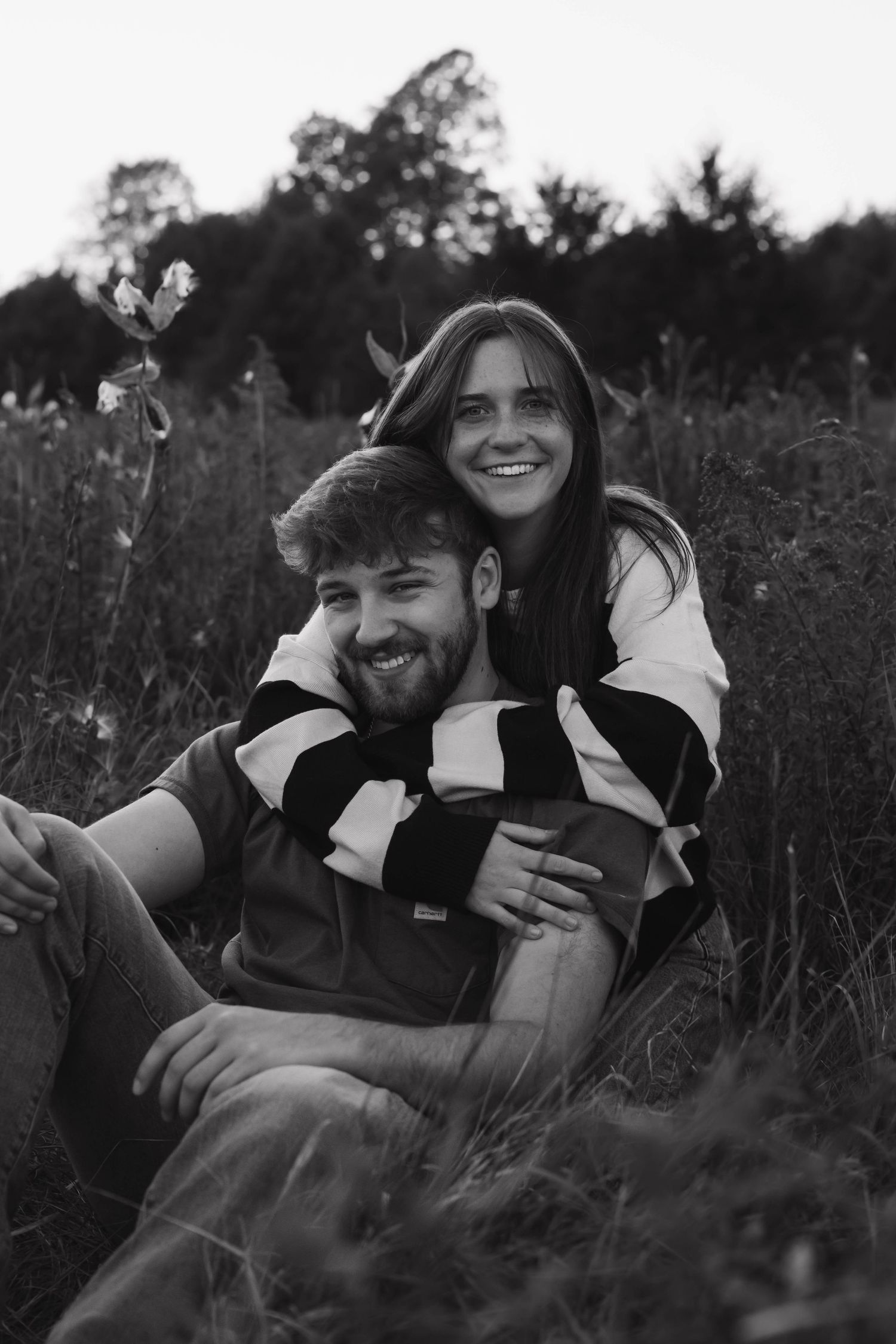Black and white photo of a smiling couple sitting in a field of wildflowers.