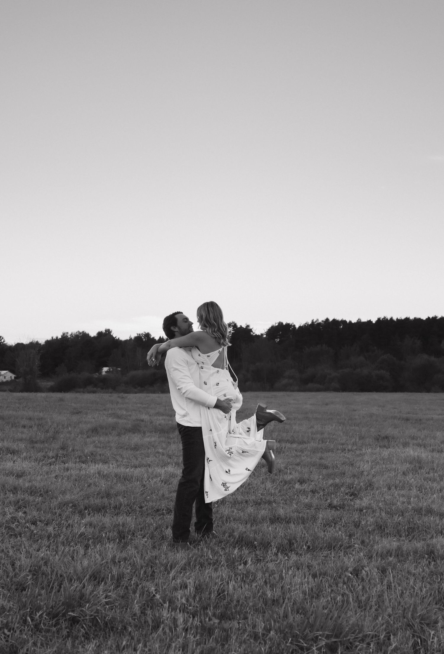 Black and white photo of a couple embracing in an open field, man lifting woman as they share a romantic kiss.