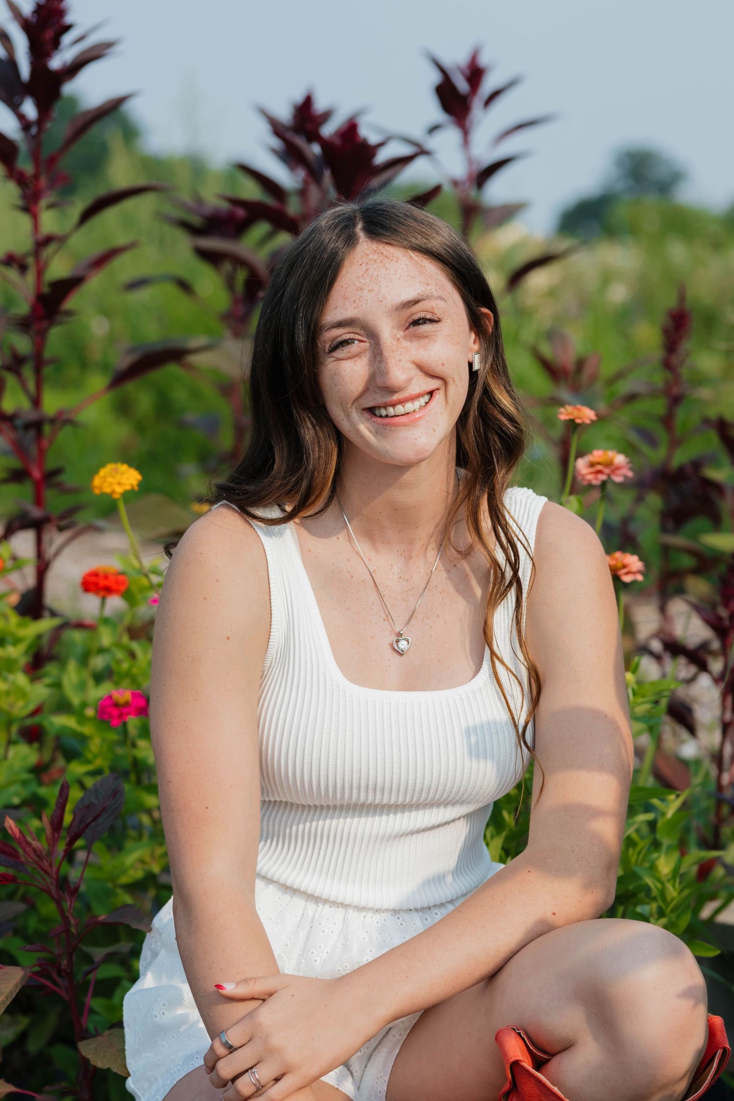 Happy teenage girl with freckles in white tank top sitting among vibrant garden flowers outdoors.