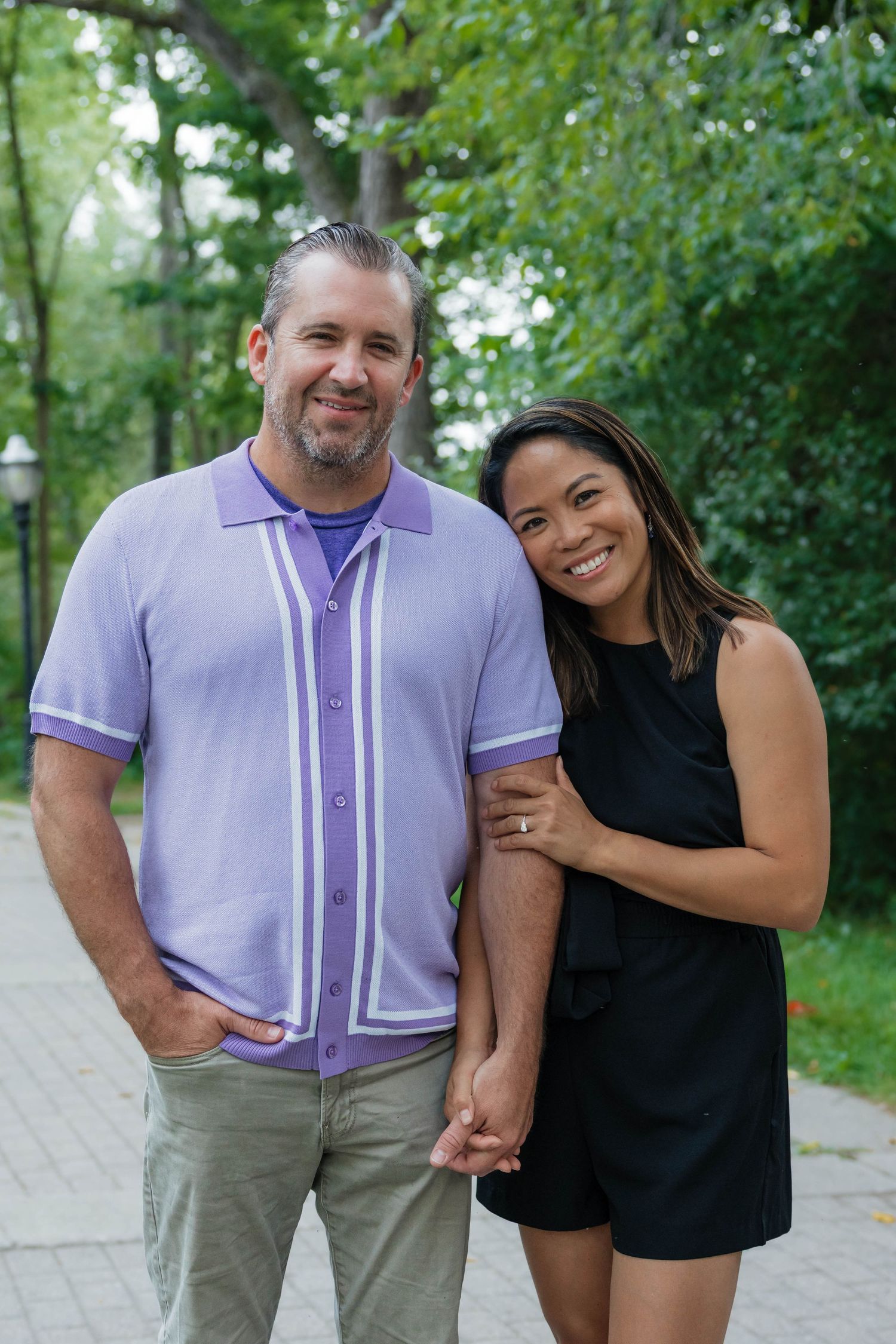 Happy couple holding hands and smiling on a tree-lined outdoor pathway.