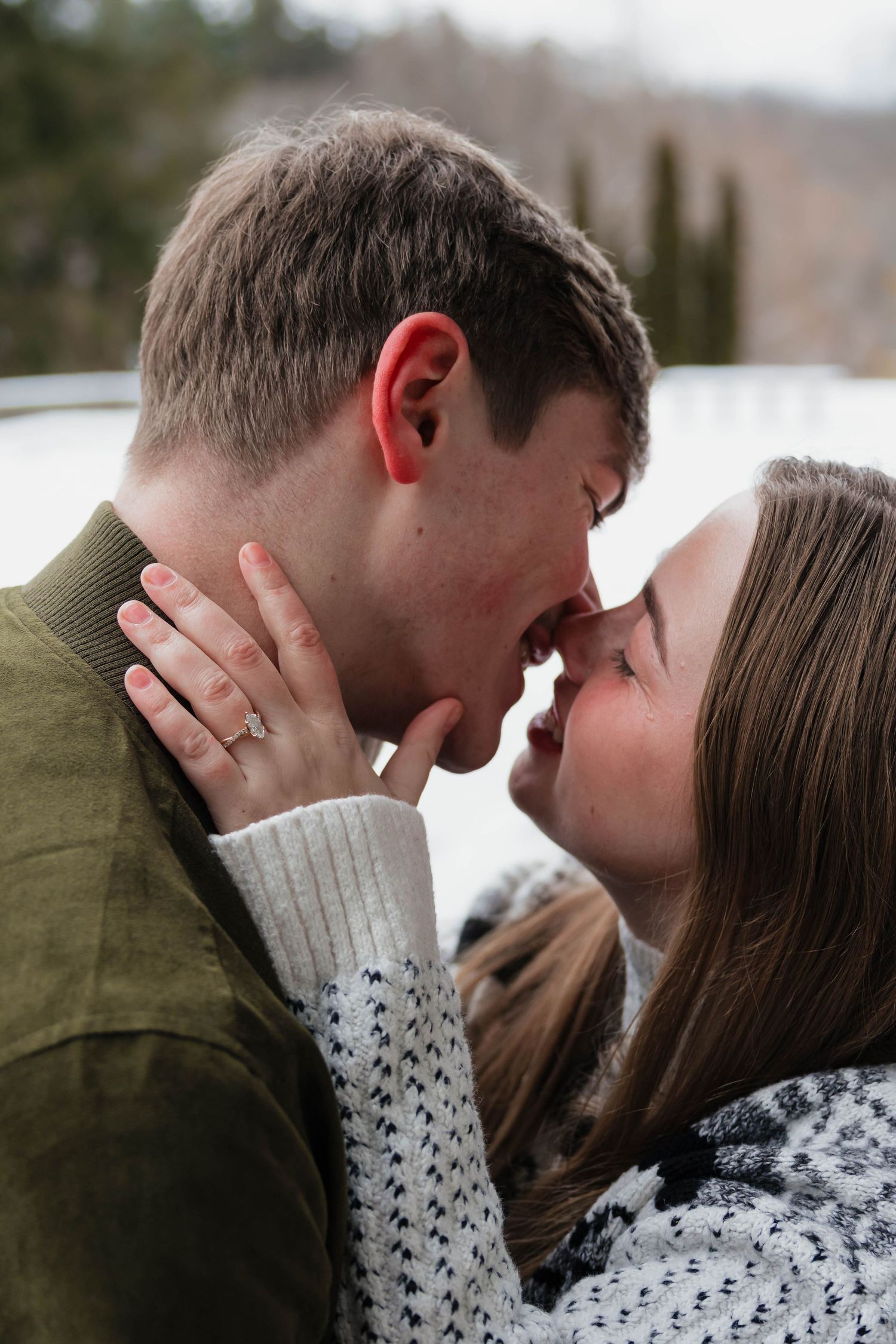 Close-up of an engaged couple about to kiss, woman showing diamond ring in a snowy setting.