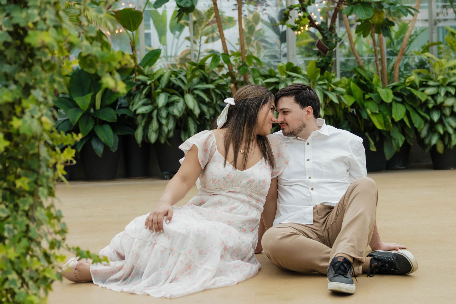 Engaged couple sitting on floor laughing together surrounded by tropical plants in a greenhouse.