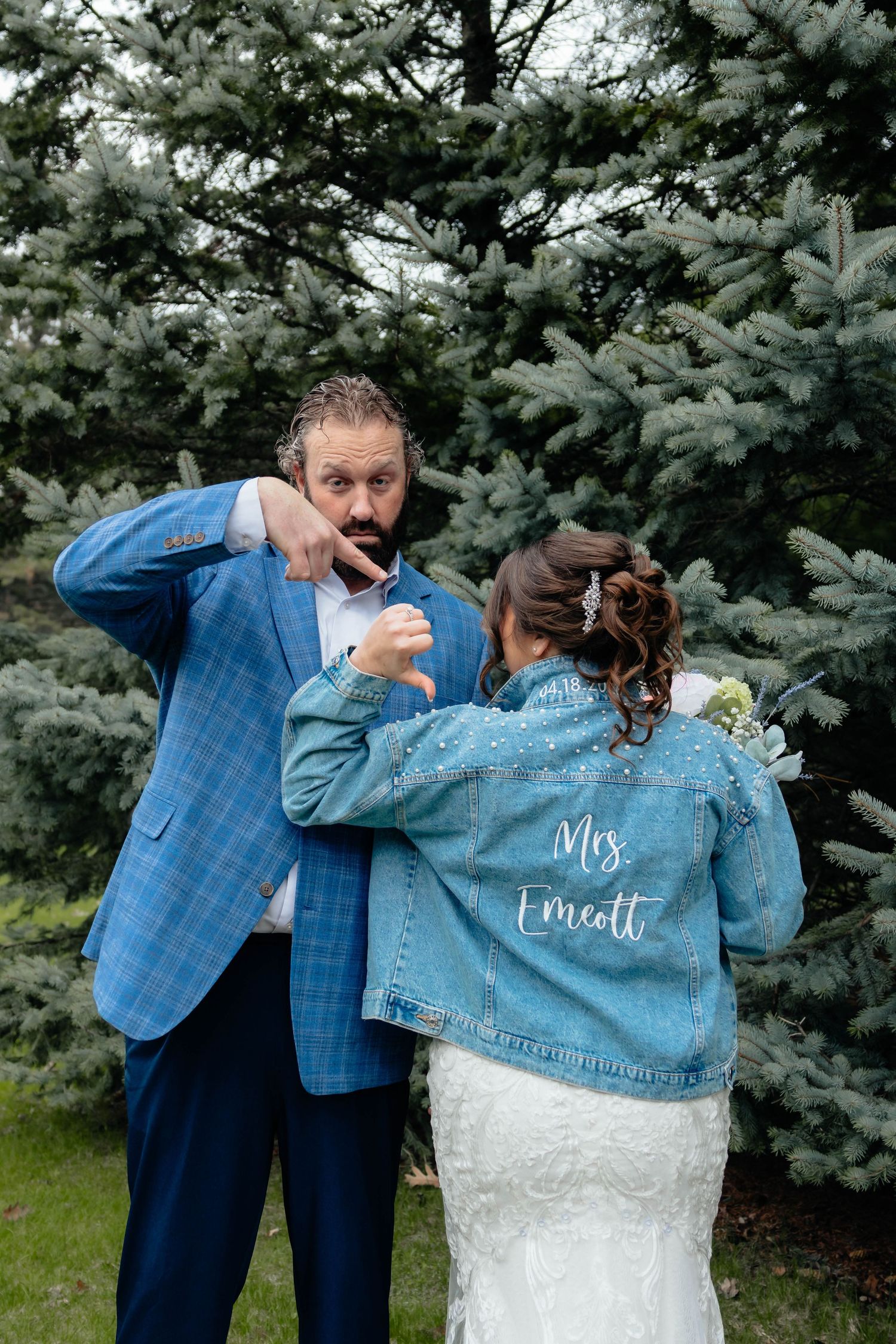 Bride in custom Mrs. Emcott denim jacket laughs with bearded groom in blue plaid suit outdoors.