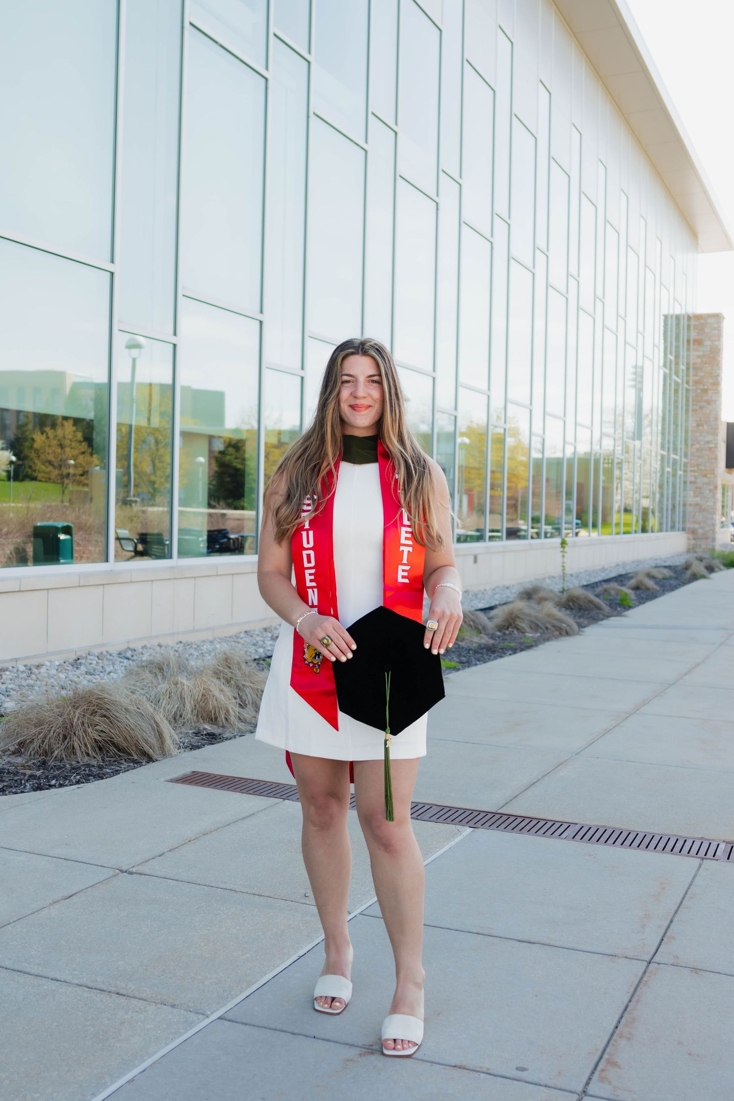 Young woman in white dress holding graduation cap and red stole outside modern glass building.