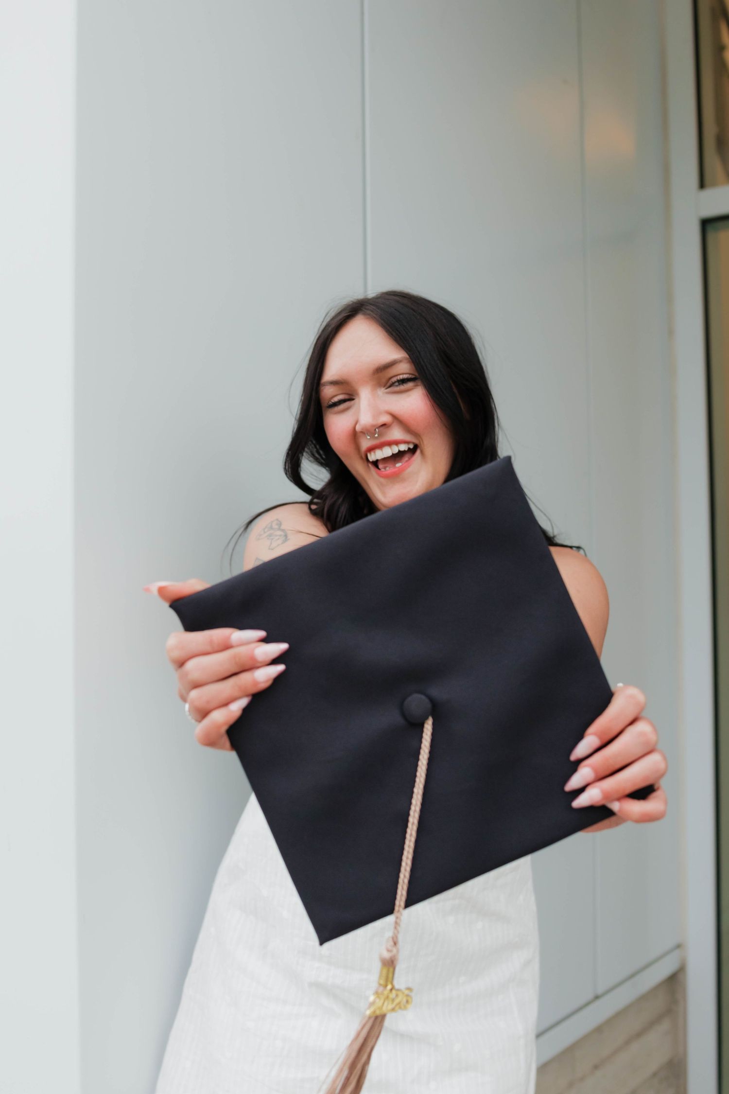 Laughing brunette graduate in white dress playfully hiding face behind black mortarboard cap.