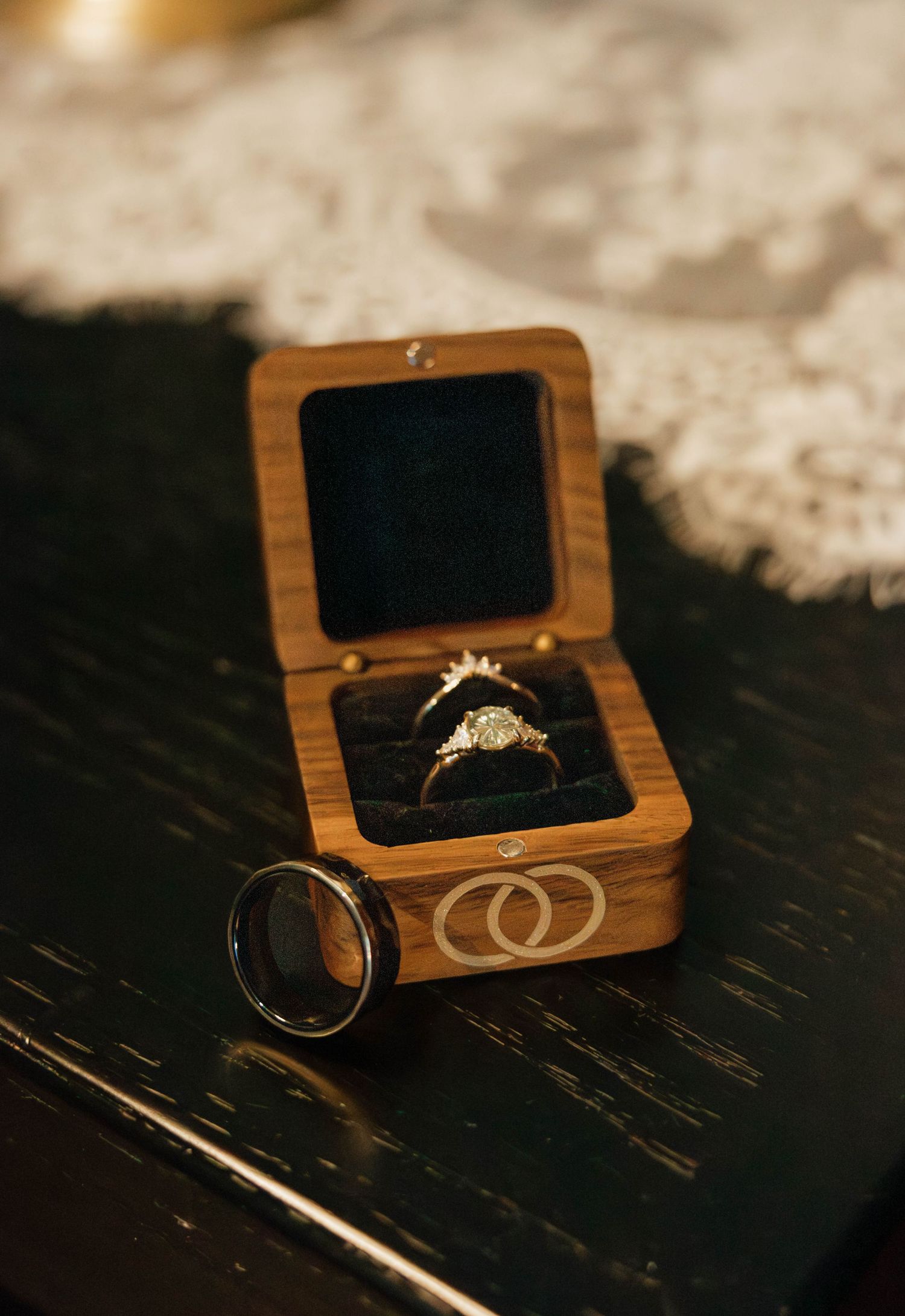 Diamond engagement ring displayed in a wooden ring box with a black tungsten wedding band beside it.