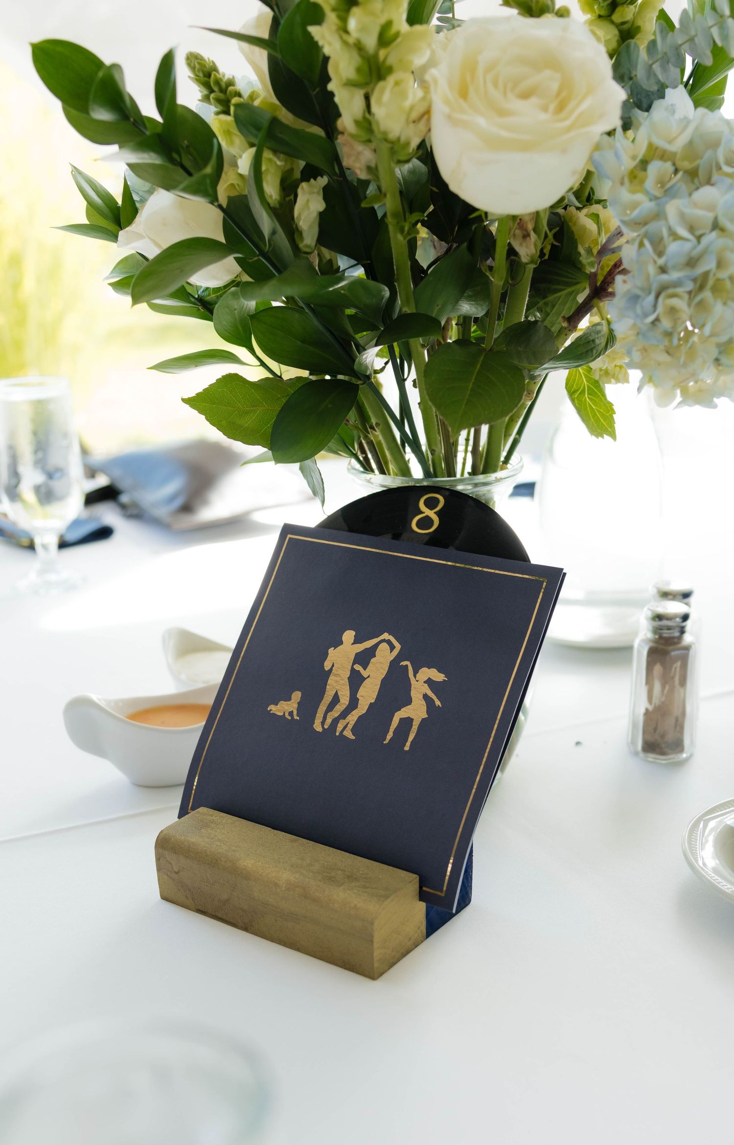 Navy and gold wedding table number card featuring dancing silhouettes in wooden holder with floral centerpiece.