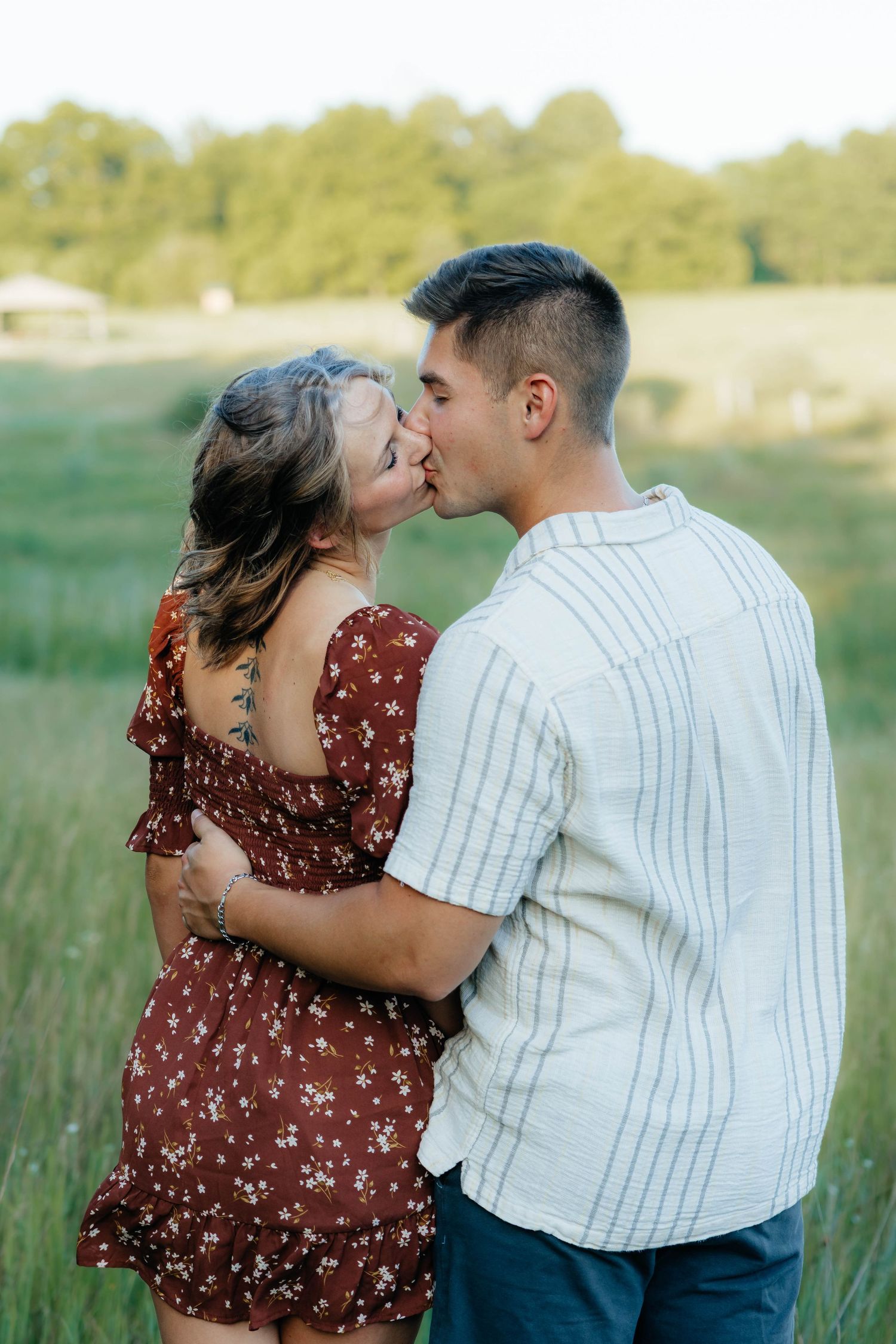 Couple sharing a kiss in an open green field, woman in floral dress with visible tattoo, man in striped shirt.