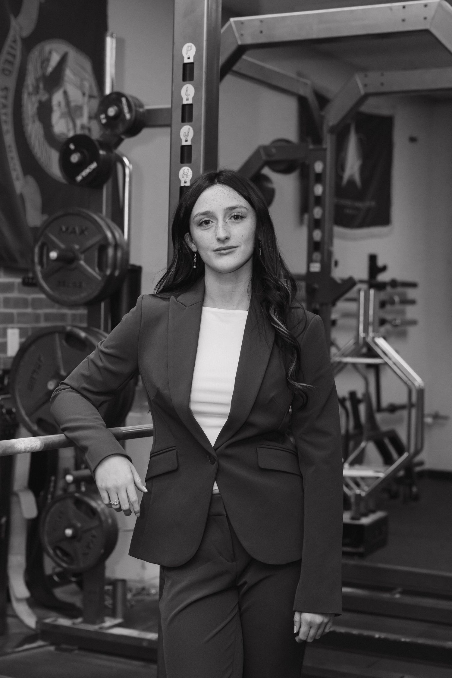Black and white portrait of a woman in a blazer posing confidently in a gym with military flags.