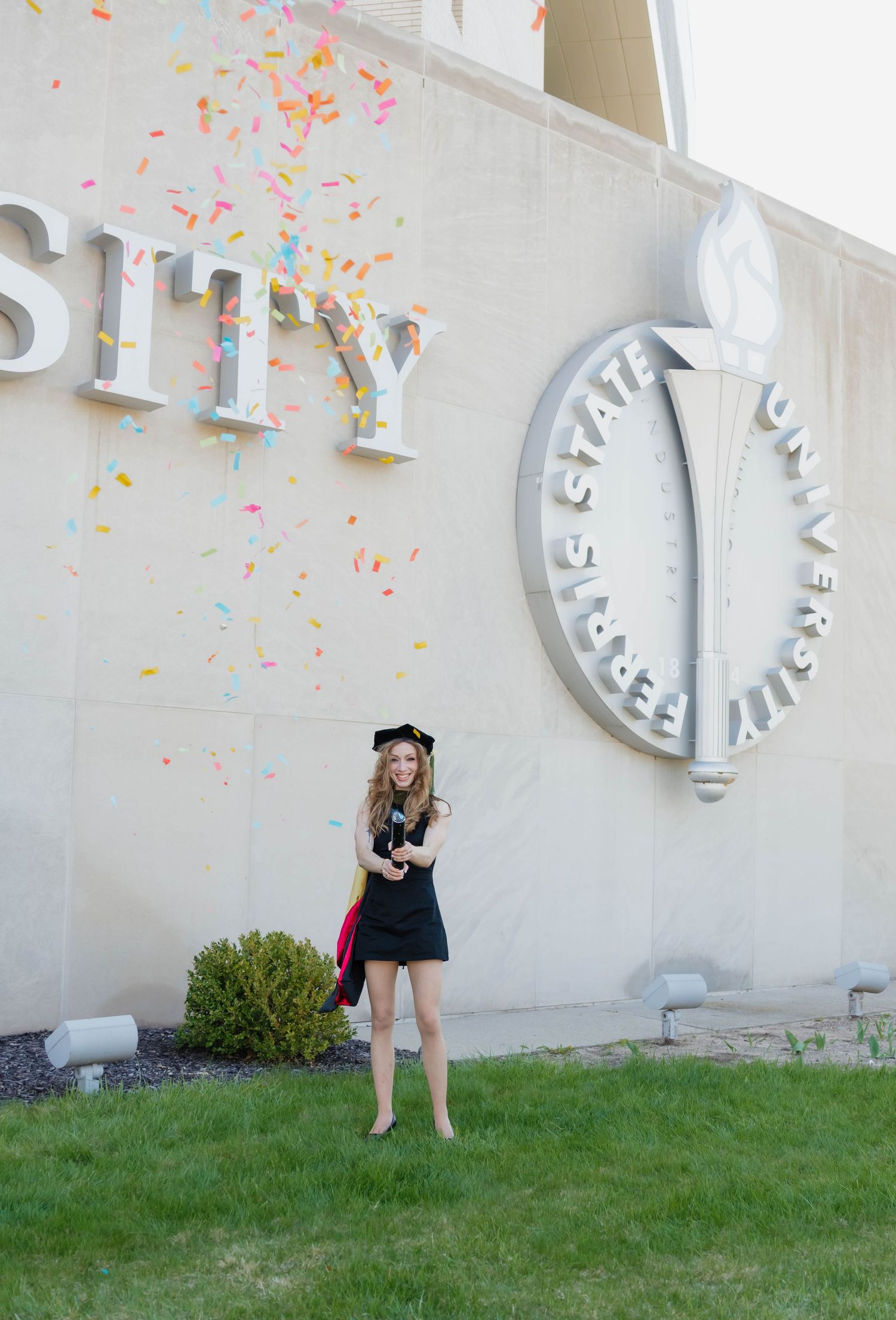 Happy graduate tossing confetti in front of Ferris State University sign on campus.