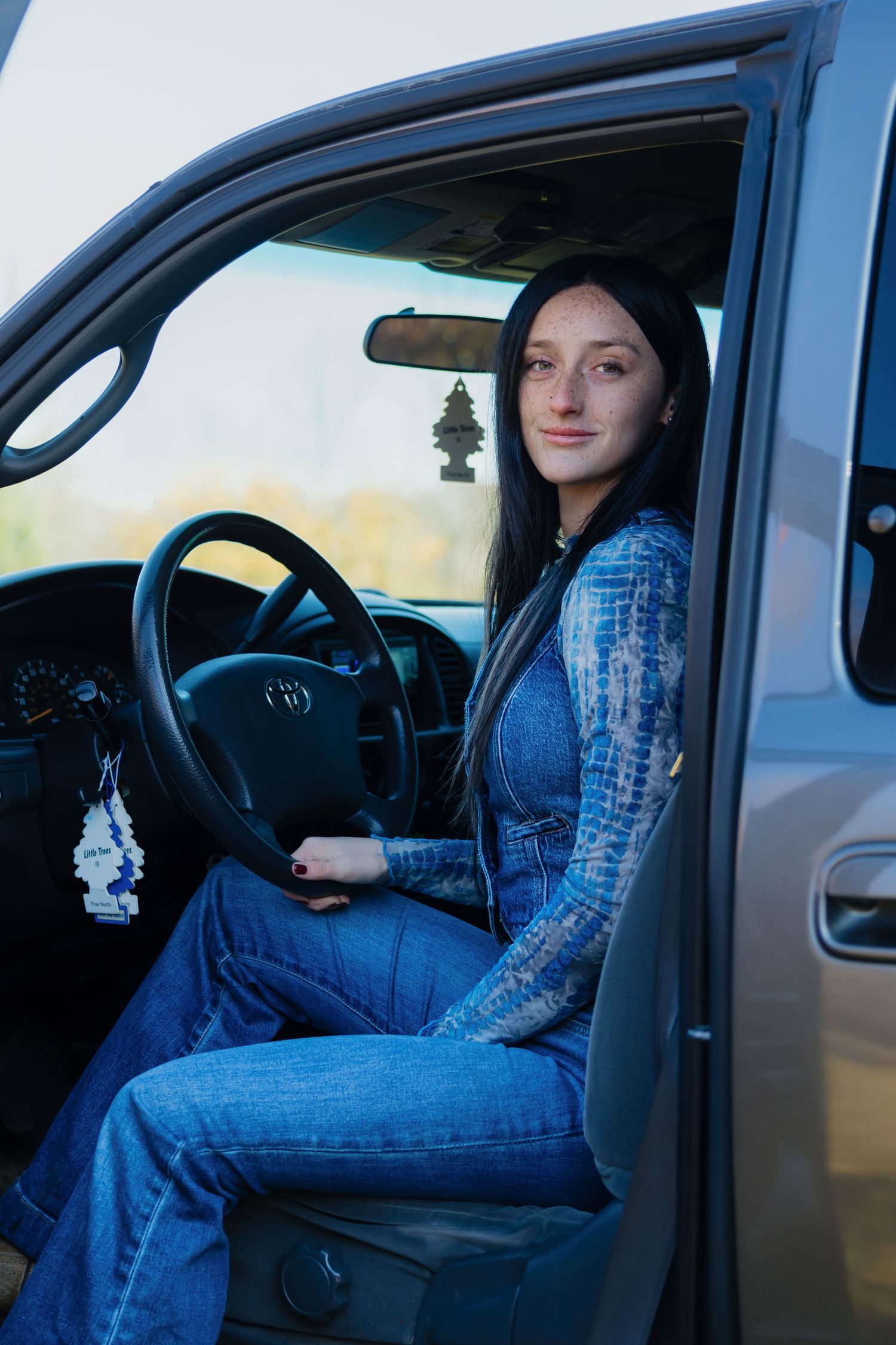 Young woman with long dark hair sitting in driver seat of Toyota truck wearing blue tie-dye top and jeans.